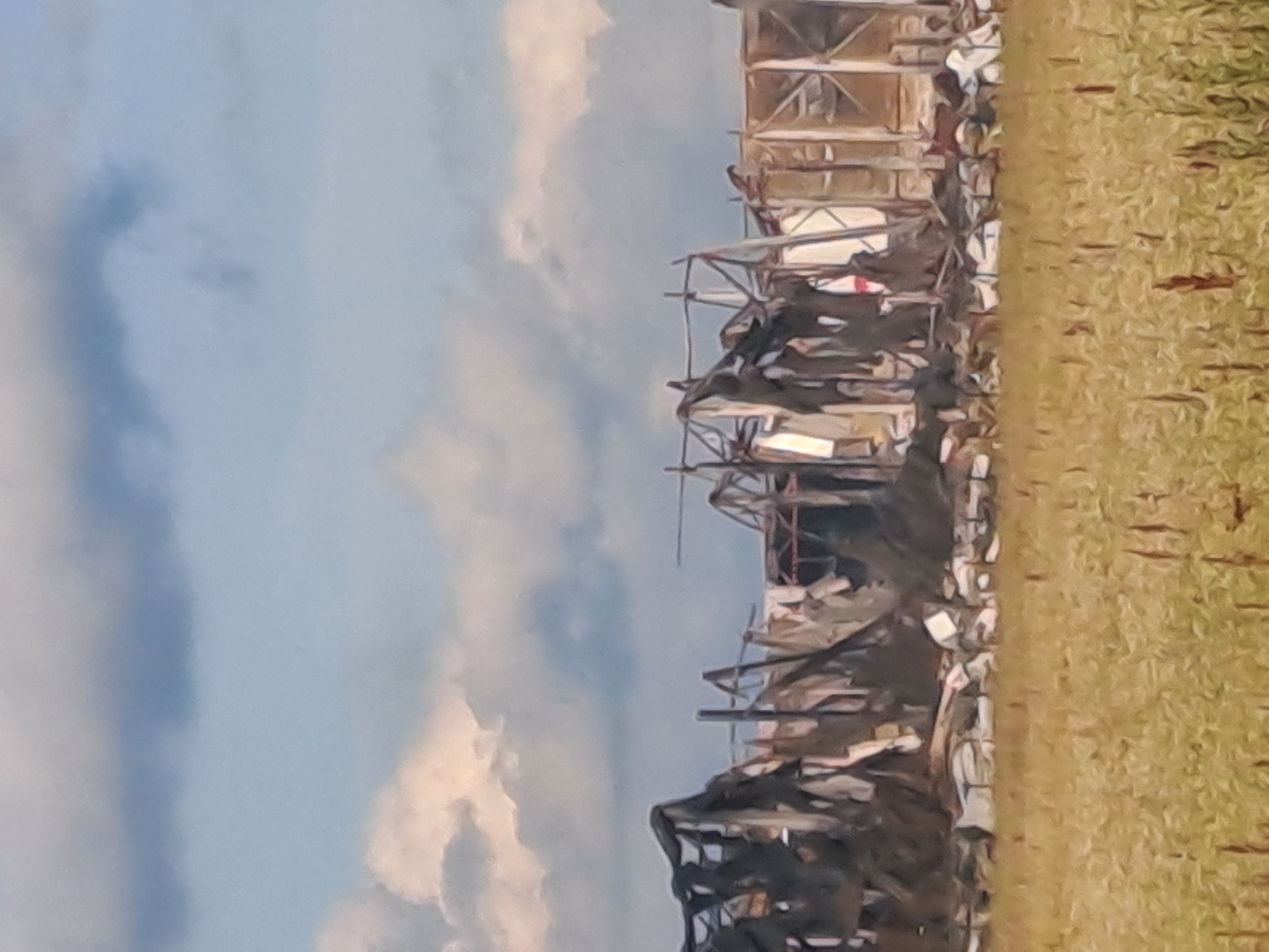 A wide shot captures a damaged building in an outdoor setting. The building, which appears to be a long structure with an open frame, shows signs of extensive damage and partial collapse. The roof is partially caved in, and some sections of the walls are missing or torn. Debris is scattered around the base of the structure. In front of the building is a field of dry, tall grass, suggesting a rural or undeveloped area. The sky above is partly cloudy, with a mix of dark and light gray clouds against a pale blue background. The overall impression is one of destruction and neglect.