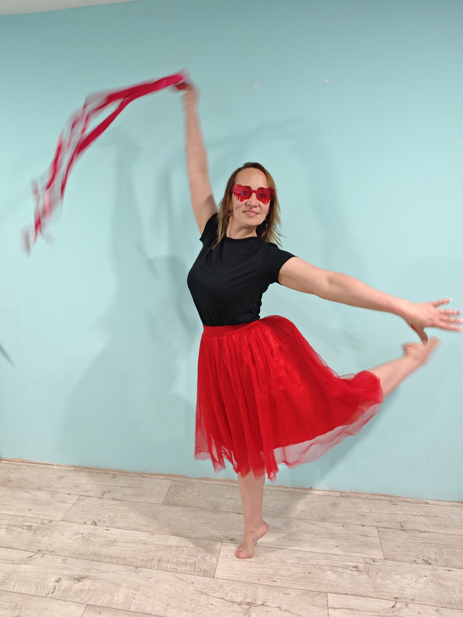 A woman stands barefoot in a dance pose in front of a light blue wall and a light wood floor. She wears heart-shaped red sunglasses, a black t-shirt, and a red tutu. She is smiling. She has one arm raised in the air holding a red and white ribbon that is blurred due to motion. The other arm is extended to the side, and her leg is raised behind her. The location is likely indoors, but the city of Markovo, Russia, cannot be verified from the image.