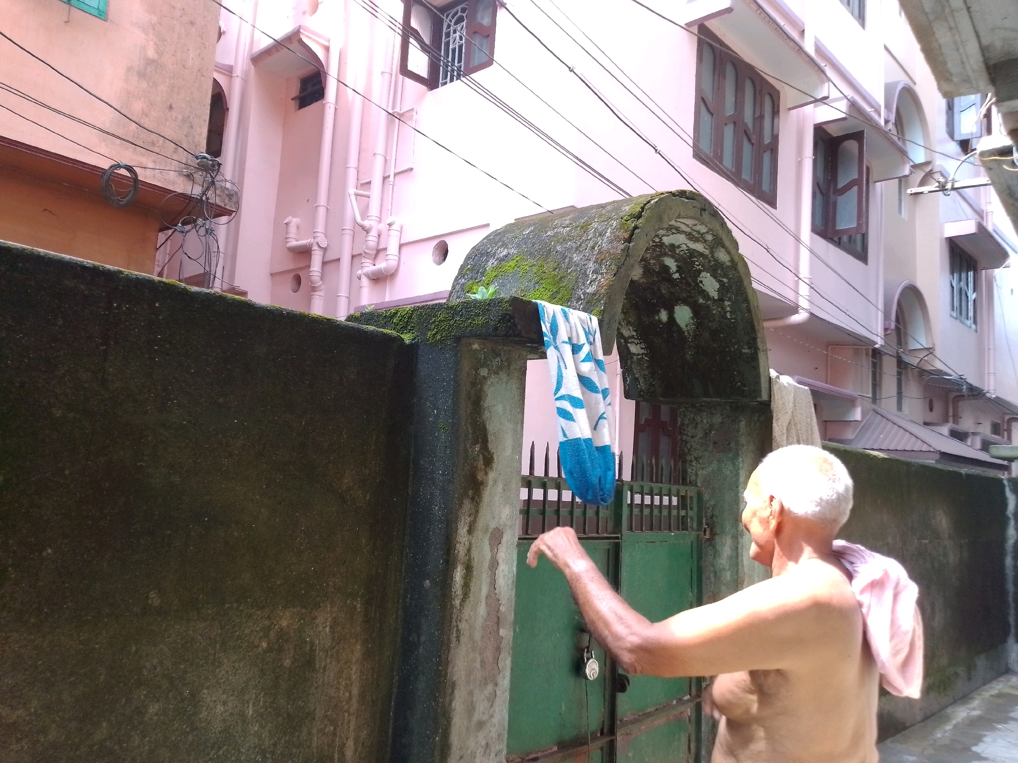 The image depicts an outdoor scene, likely in a residential area of Siliguri, India, given the architectural style and the presence of overhead wires common in Indian cities. In the foreground, a man, appearing to be middle-aged or older with short white hair and bare chest, is partially visible. He is facing away from the camera, towards the right side of the frame, with his left arm extended. A pink towel or cloth is draped over his right shoulder and back.
Behind the man is a green metal gate, which is locked with a silver padlock. Above the gate, a blue and white patterned towel hangs. The gate is set against a weathered, moss-covered wall that extends into the left side of the image. Beyond the wall and gate, a light pink building with several windows and visible pipes runs along the background. Overhead, a network of black electrical wires crisscrosses the scene, a characteristic feature of urban India. The overall lighting suggests it is daytime, with the sun casting shadows and illuminating the man from the side. The environment appears somewhat cluttered and utilitarian, common in many Indian urban settings.