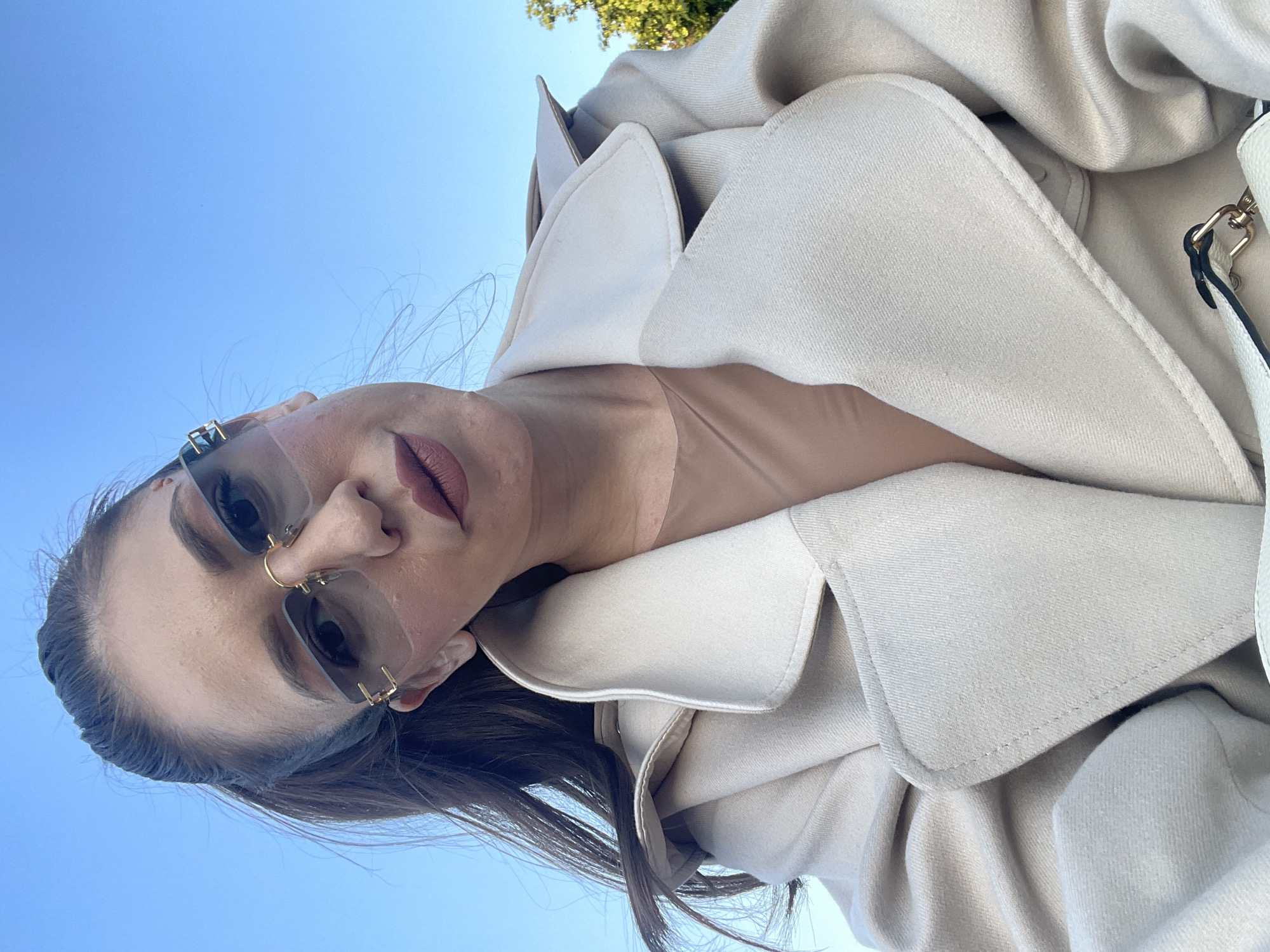 A medium shot depicts a woman with light brown hair and tan skin, wearing gold-framed sunglasses with nose jewelry, angled in a way that the sky is visible in the background. She is wearing a light beige overcoat, likely made of wool or a similar heavy fabric. The location is not discernible from the image. Bulgaria, or the city of Sofia, cannot be determined from the photograph.