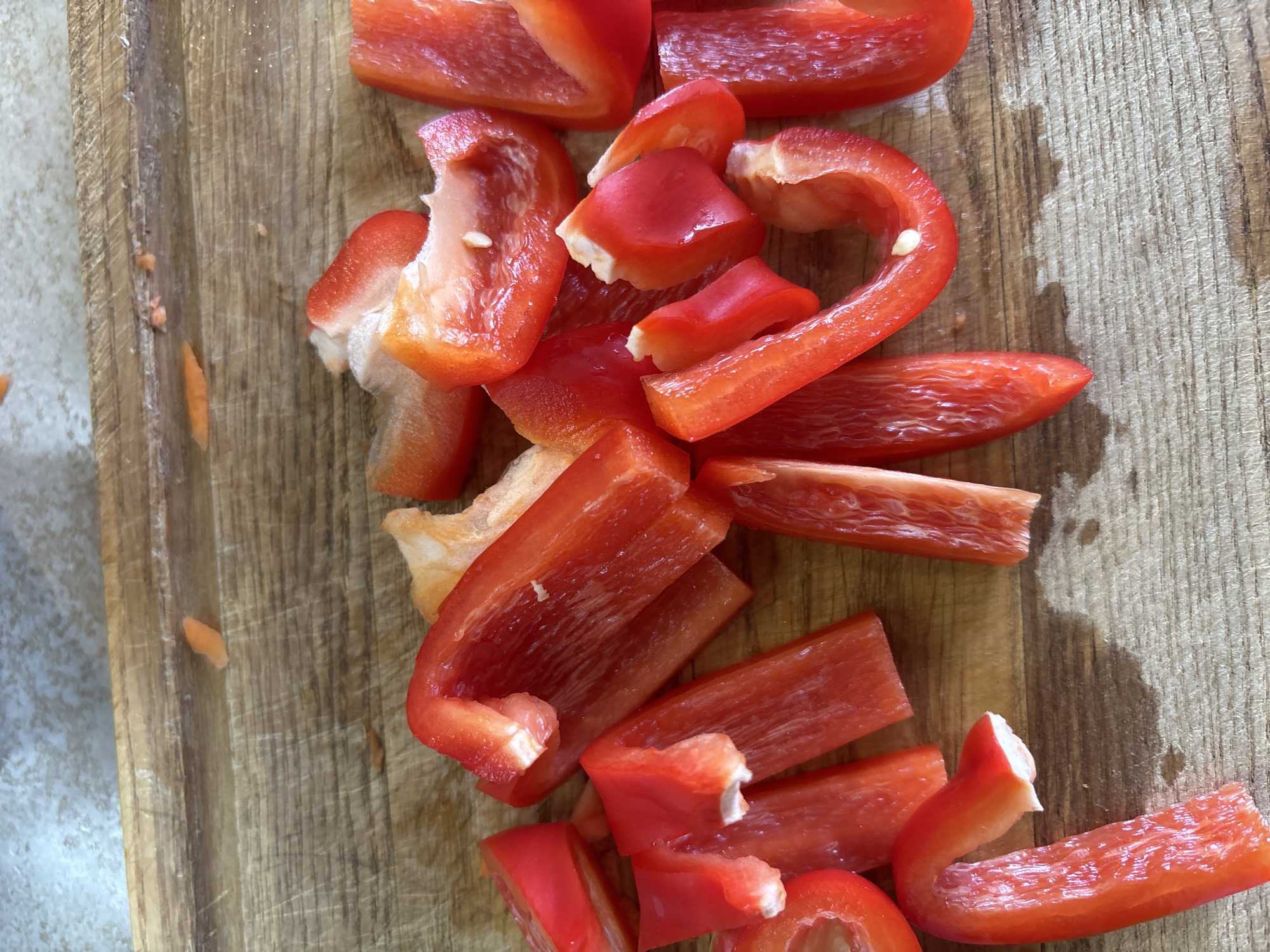 The image shows a pile of chopped red bell peppers on a wooden cutting board. The peppers are cut into various shapes and sizes, including strips, chunks, and curved pieces. Some of the peppers still have seeds attached. The wooden cutting board has a visible grain and appears to be well-used, with some scratches and water stains. In the background, a light-colored, possibly marble or granite surface is visible, with some small orange specks, potentially bits of carrot or other vegetables. The lighting suggests an indoor setting, possibly a kitchen. The overall impression is one of food preparation. No people, text, time of day, or weather cues are discernible. The location context is Bila Tserkva, Ukraine.