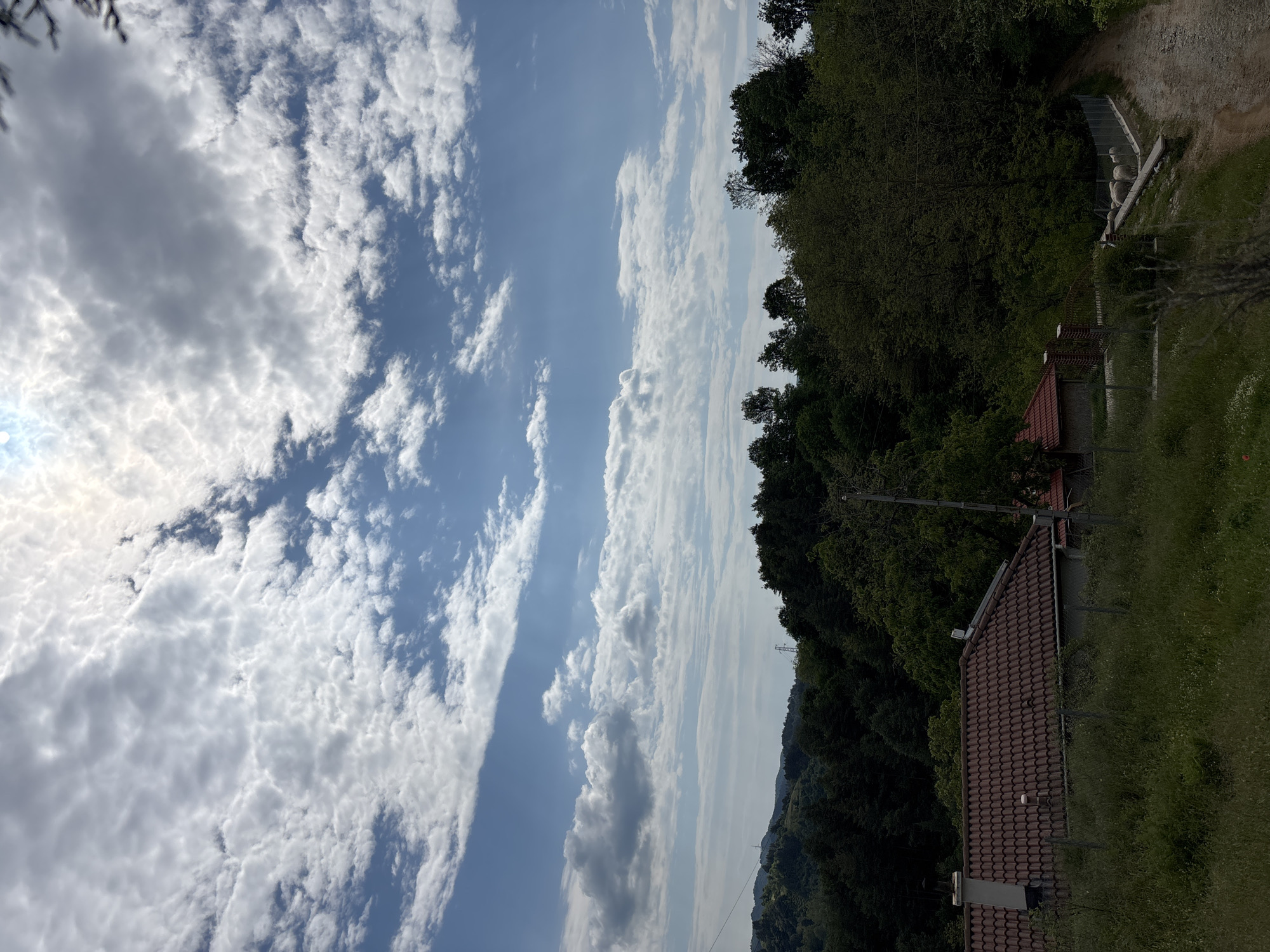 The image depicts a daytime outdoor scene in Zlatograd, Bulgaria. The sky is predominantly blue with extensive cloud cover, featuring both small, puffy clouds and larger, elongated formations. A line of dense, dark green trees covers the right side of the frame, descending from the top edge and extending down the side. At the base of the tree line, a building with a brown tile roof is visible. A grassy area is near the bottom of the frame. A dirt path can be seen behind the trees.