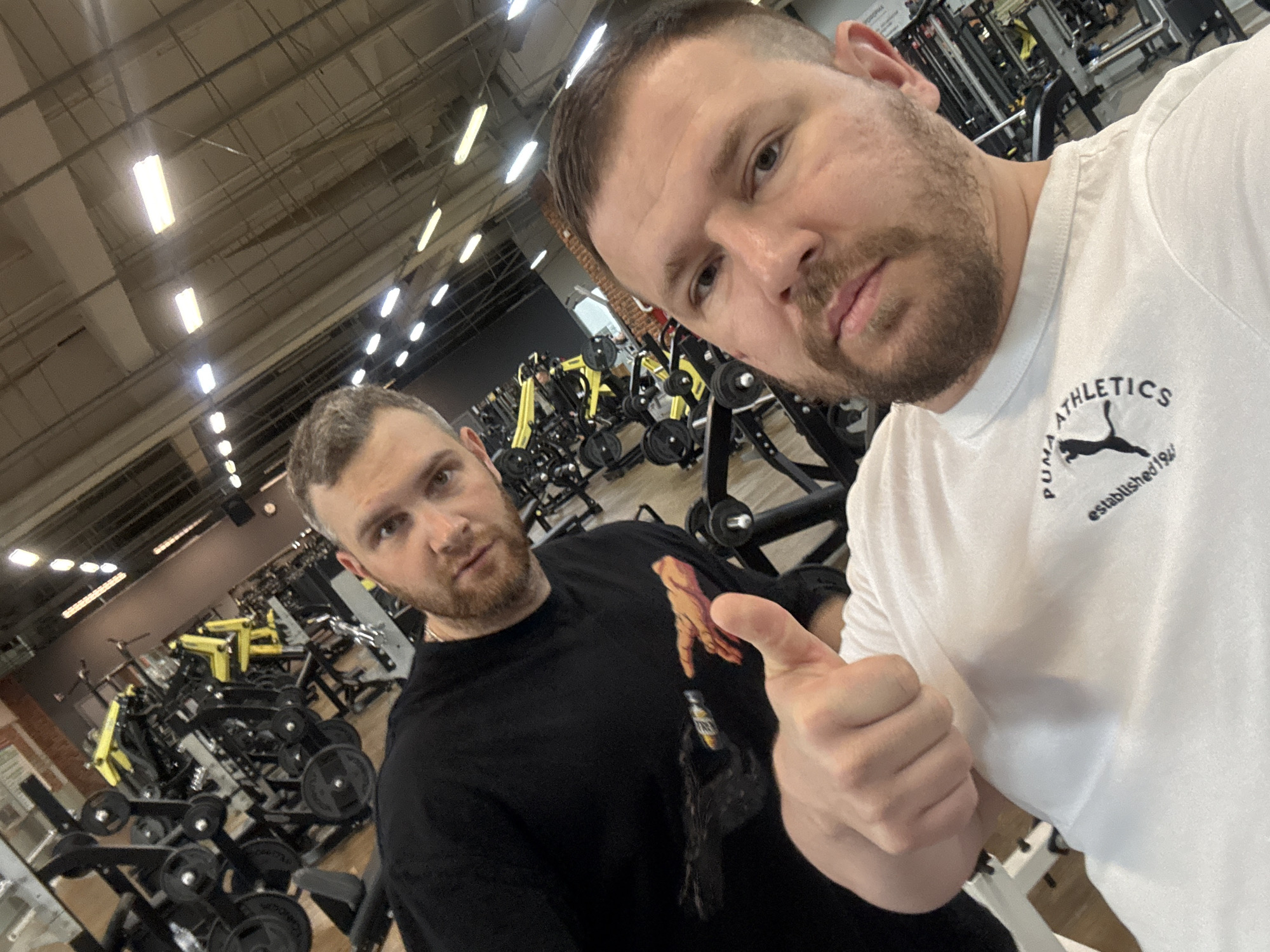 The image shows two men in a gym in Chaikovsky, Russia. The gym has weight training equipment in the background, with yellow and black colors. The man in the foreground wears a white t-shirt with Puma Athletics branding. He is giving a thumbs up. The other man wears a black t-shirt. The lighting in the gym is provided by overhead fluorescent lights.