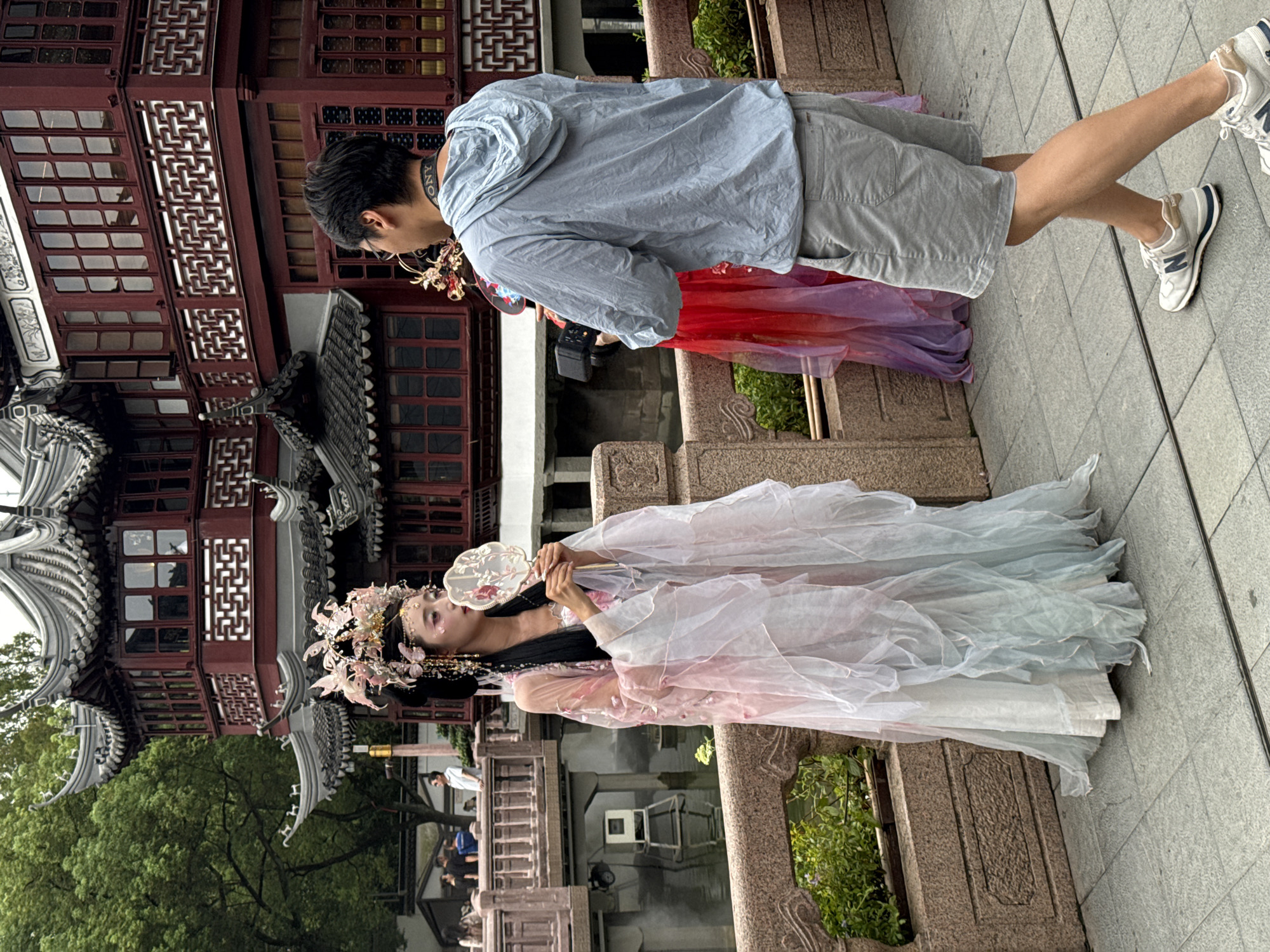 A woman in traditional Chinese attire and a headdress stands on a stone bridge, holding a fan. She is posing for a man who is filming her with a video camera. In the background, a traditional Chinese building with red walls and ornate roofs is visible, along with lush green trees. Another person in a red and purple dress is visible behind the man. The scene appears to be set in a garden or park, likely in Shanghai, China.