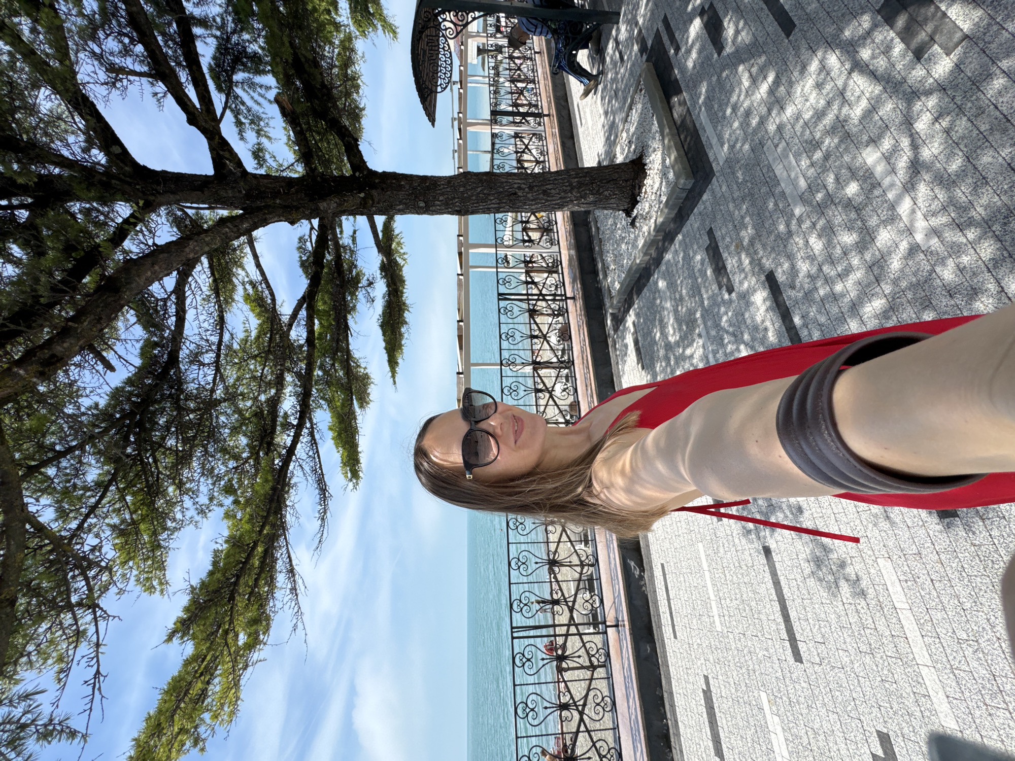 This image, set in Sochi, Russia, depicts a woman taking a selfie on a sunlit promenade adjacent to the Black Sea. The woman, positioned in the lower-right foreground, has light brown hair, wears dark sunglasses and a red dress, and is smiling towards the camera held by her outstretched right arm. The promenade is paved with light and dark gray rectangular tiles, patterned with shadows cast by the dense foliage of a large tree. The tree, located centrally in the mid-ground, features a thick, dark trunk within a low, white-pebbled planter. To the right of the tree, a man is seated on an ornate black metal bench with a curved canopy, facing away from the camera. A decorative black metal fence with heart-shaped scrollwork separates the promenade from a narrow, light-colored beach. Beyond the beach, several white open-frame structures are visible along the coastline. The background features the expansive, tranquil turquoise waters of the Black Sea beneath a light blue sky with wispy white clouds.