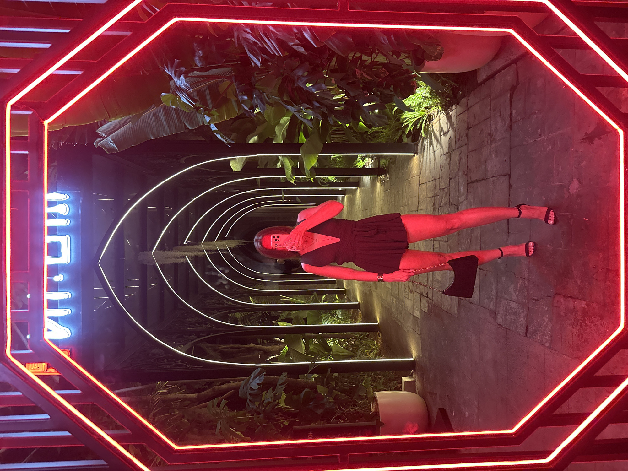 A woman is standing in a walkway lined with lush greenery and illuminated by glowing white arches that create a tunnel effect. She is facing a large octagonal frame with bright red neon lights. The woman is holding a mobile phone in her right hand, seemingly taking a mirror selfie. She is wearing a black V-neck romper and black strappy high heels. A small black shoulder bag with a gold chain is hanging from her left shoulder. The walkway appears to be paved with stone. In the background, a neon sign is visible at the end of the tunnel, with lettering that is difficult to decipher. This scene is located in the city of North Kuta, Indonesia, likely at a venue with an emphasis on dramatic lighting and exotic plant life.