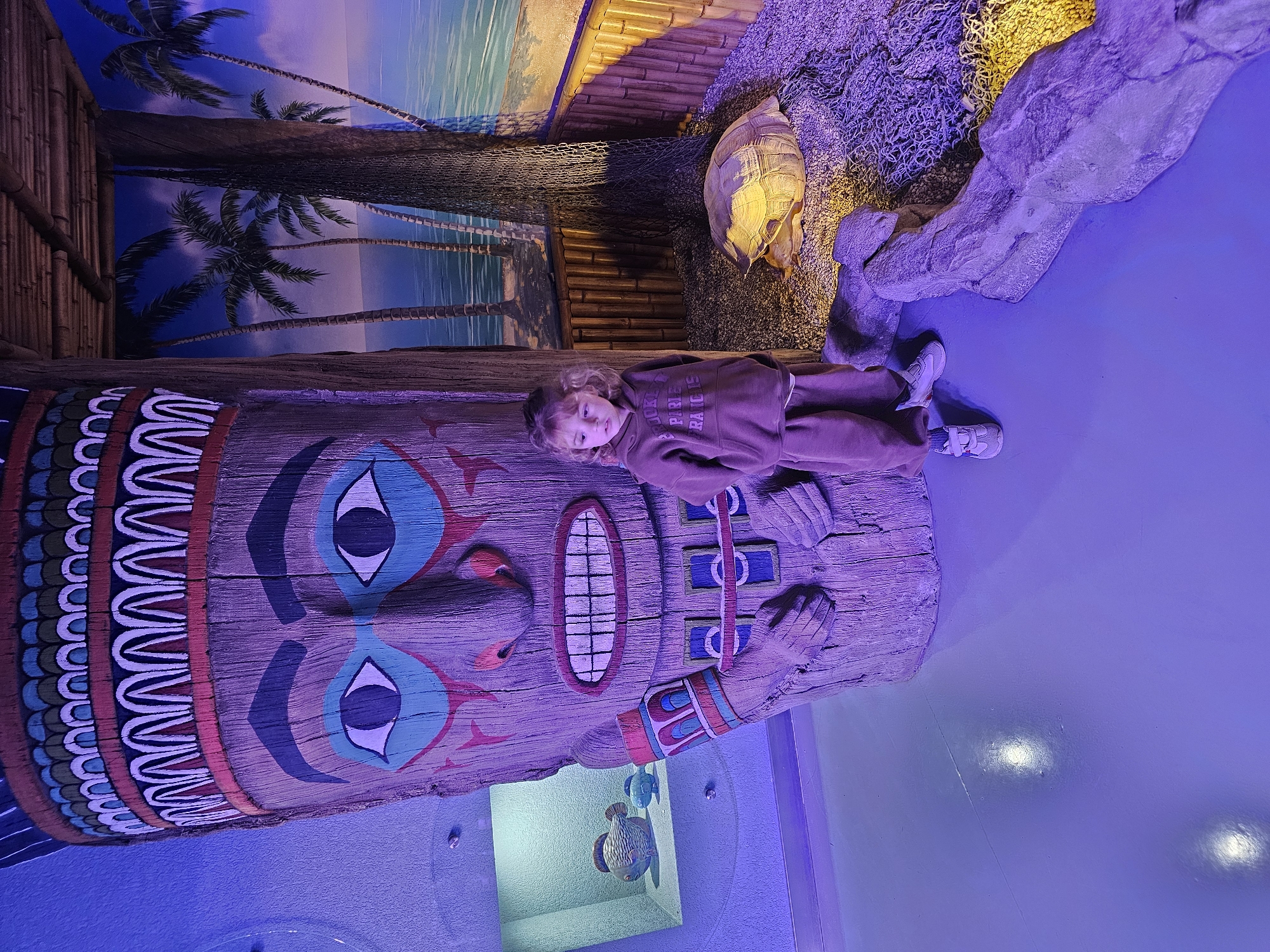 A color photograph depicts a child lying on a large, carved wooden totem pole. The totem pole features painted details including eyes, a mouth with teeth, and patterns. The child is wearing a brown sweatshirt and pants and is positioned lengthwise on the totem pole, looking toward the viewer.
The background consists of a beach-themed mural, bamboo structures, netting, and a turtle figure. A faux rocky outcrop extends onto a blue-colored floor, which is likely part of an indoor exhibit. A decorative fish aquarium is visible in the lower right corner.
The image was taken at the Aquarium in the city of Khimki, Russia.