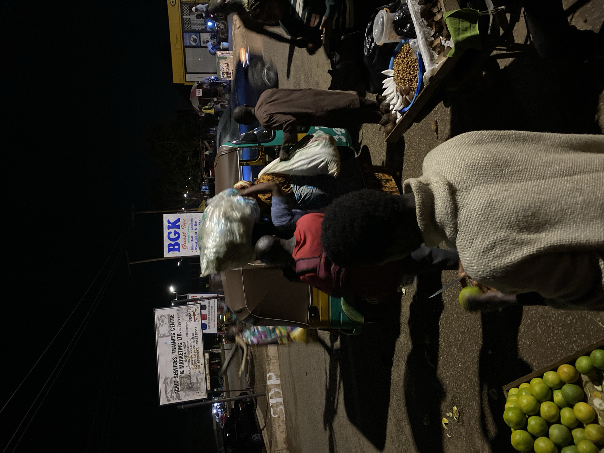 This nighttime image captures a bustling street market scene in Bukuru, Nigeria. The setting is illuminated by artificial streetlights, casting shadows of people and objects on the asphalt road.
In the foreground, to the bottom right, a person wearing a light-colored, possibly knitted, garment is actively cutting a green fruit, likely an orange or lime. Adjacent to them is a wooden crate filled with an abundance of similar green and yellow fruits. Just behind this activity, a roadside stall or vendor's display offers various items, including what appear to be seeds or grains in a blue bowl, white peeled fruit (possibly bananas), and other produce.
The middle ground is dominated by a green and yellow tricycle (tuk-tuk), around which several individuals are gathered. One person in a red shirt is seated on the tricycle, while another in a brown robe stands nearby. Plastic bags, some filled with goods, are visible, suggesting transactions or transport of merchandise.
The background reveals more activity with people and vehicles, and structures including a building with a yellow facade. Overhead power lines crisscross against the dark sky. Visible text on signs includes "AGRIC SERVICES, TRAINING CENTRE & MARKETING LTD. RC: 98149" with contact details such as email, website (www.astc.ng), and phone numbers, and a postal address for "JOS, PLATEAU." Another sign on the yellow building reads "BGK Guest Inn," advertising "Mini Hall," "Challets" (likely chalets), and a "Resturant & Bar" (likely restaurant), and mentions "Kwala Zawan." The word "SDP" is also painted on the road surface, and "VOM" appears on a smaller sign. The overall scene depicts a vibrant commercial area after dark.
