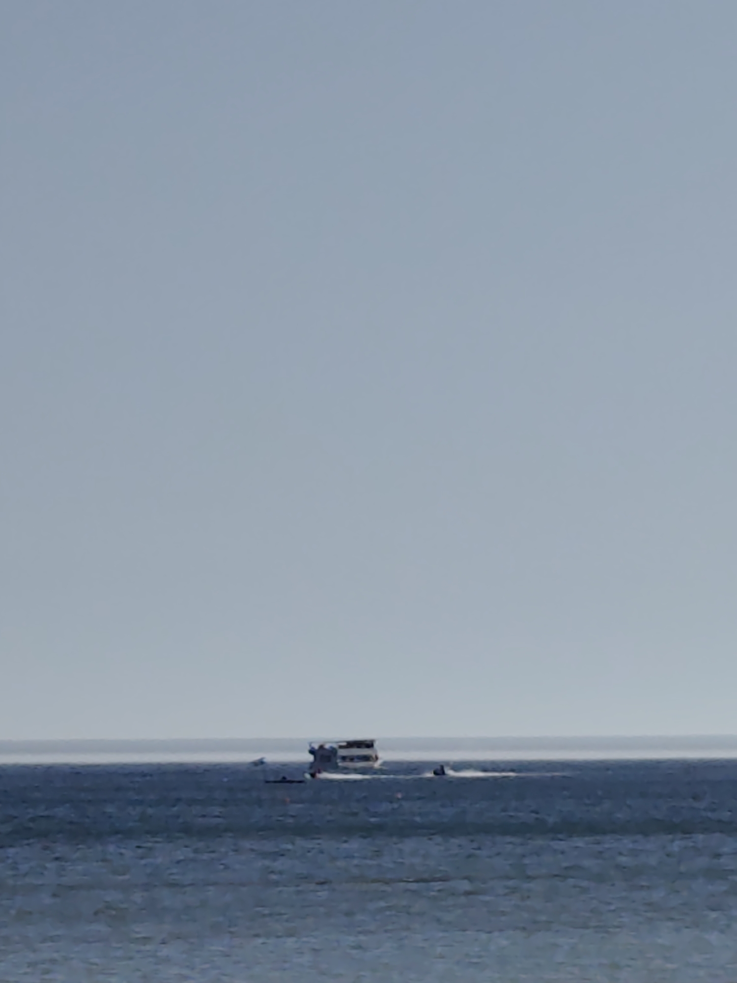 A wide-angle, low-angle shot captures a calm body of water, likely the Caspian Sea near Aktau, Kazakhstan. The horizon line is sharply defined, separating the deep blue water from a pale, hazy sky. In the middle distance, a white passenger boat with multiple decks is cruising on the water. To the left of the passenger boat, a smaller black vessel, possibly a jet ski or a small boat, is visible. The water surface is relatively smooth with small ripples. The overall impression is one of a serene, open-water scene on a clear day.