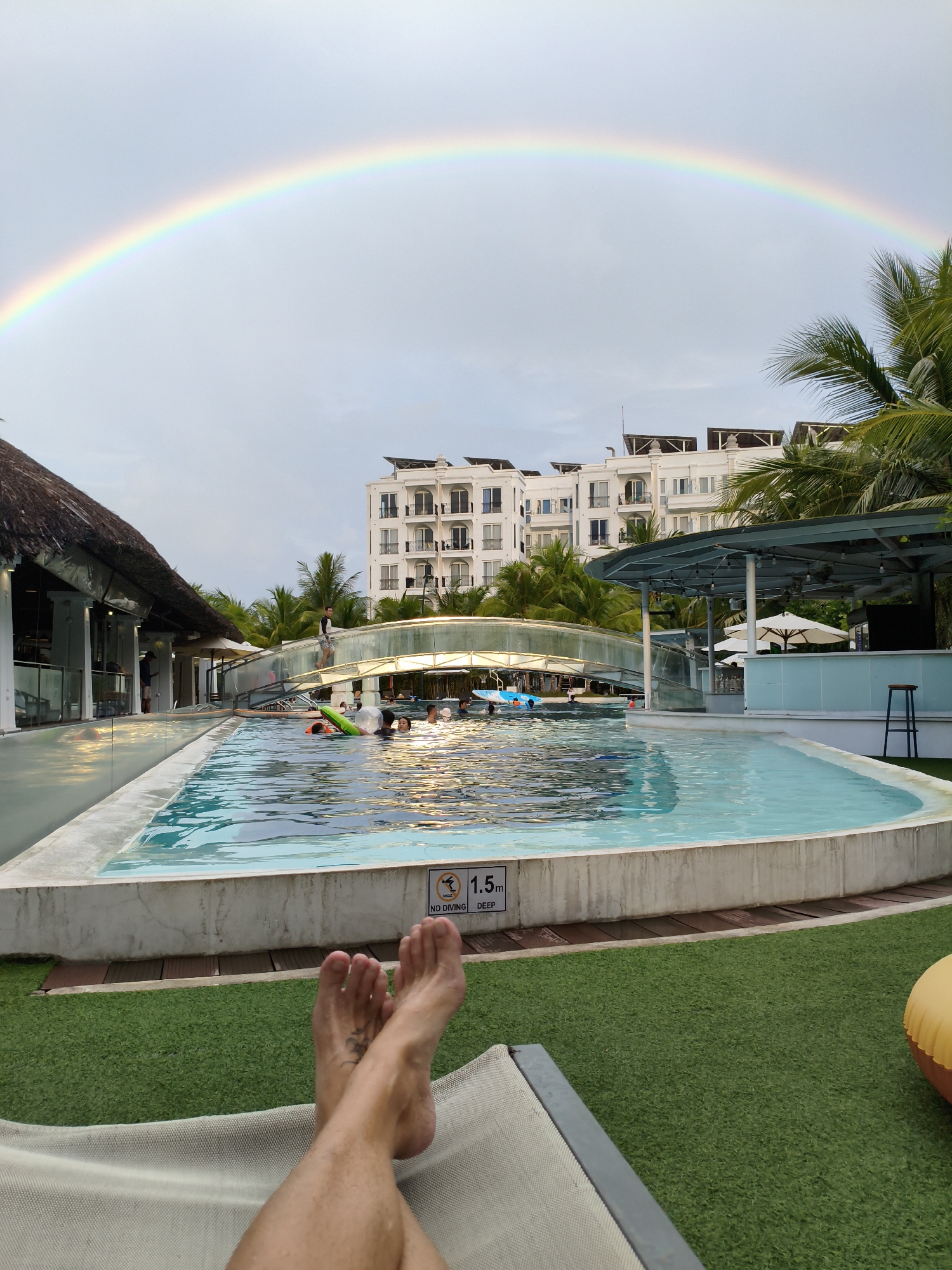 A wide-angle, eye-level shot captures a resort swimming pool area in Nha Trang, Vietnam, under a sky adorned with a vibrant rainbow. In the foreground, a person's bare feet are crossed over a recliner, positioned on a patch of artificial grass. The pool itself, with its bright turquoise water, occupies the mid-ground, with several people swimming and floating. To the left, a building with a thatched roof and open-air seating is visible. A white, multi-story building with arched windows stands in the background, surrounded by palm trees. A pedestrian bridge arches over the pool, connecting different areas of the resort. A sign indicating "1.5m DEEP" and "NO DIVING" is attached to the pool's edge.