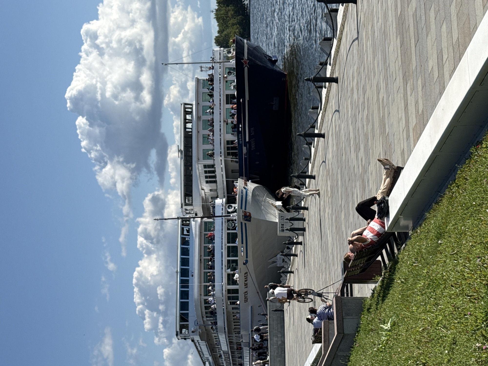The image captures a sunny day at a waterfront in Khimki, Russia, likely along the Khimki Reservoir, which is part of the Moscow Canal. In the background, two large river cruise ships are docked side-by-side. The ship on the left is white with multiple decks, labeled "СЕРГЕЙ ДЯГИЛЕВ" in blue lettering. The ship on the right is dark blue and is labeled with Russian text, including a red star near its bow. The decks of both ships are populated with passengers.
The foreground features a paved promenade along the water's edge, bordered by a railing with black posts and chain. To the left of the promenade, a grassy embankment slopes down from the walkway. On the promenade, several individuals are visible. A man on a bicycle rides towards the ships, while another person stands near the water's edge looking at their phone. Further to the left, several people are seated on benches, including two men who appear to be relaxing on a sun lounger, one wearing a striped shirt. The sky is blue with large, fluffy white clouds.