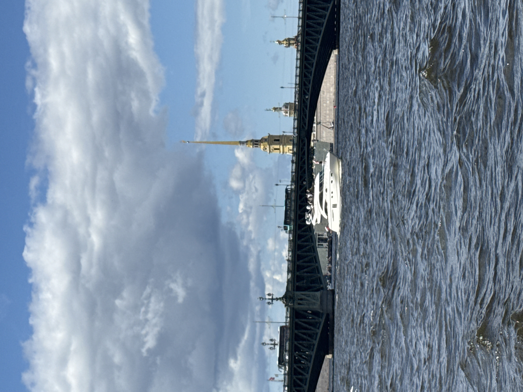 This image captures a view of the Neva River in Saint Petersburg, Russia, with the Palace Bridge prominently featured. A sleek white yacht is sailing on the river in the foreground, its reflection visible on the choppy water. The Palace Bridge, a wide, green truss bridge, spans the river, with several vehicles, including buses, traversing its surface. In the background, behind the bridge, the iconic spire of the Peter and Paul Fortress, a golden needle-like structure, pierces the cloudy sky, accompanied by other architectural elements of the fortress complex. The sky is a mix of blue patches and heavy, gray clouds, suggesting a dynamic weather system. The overall scene depicts a lively urban waterway with significant historical and architectural landmarks.