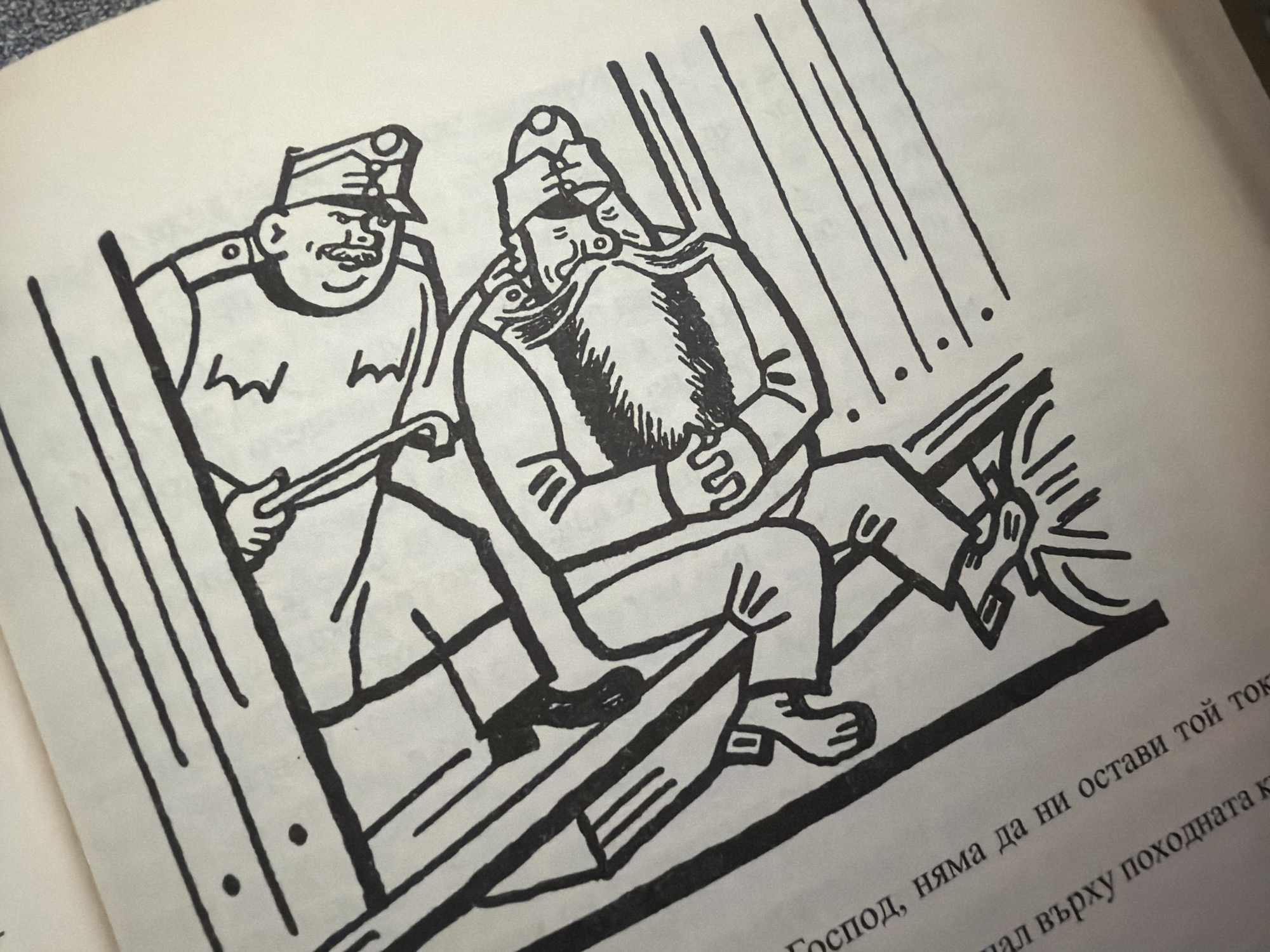 The image is a black and white line drawing of two cartoonish figures. The figures are positioned on what appears to be parallel lines, suggesting a railway or tracks. One figure is leaning over another, and there is a tool held by the leaning figure that is interacting with the lower figure. Both figures are wearing hats and appear to be men. The drawing is on a page of a book, with text visible in the lower left corner. There is no indication of the city of Pazardzhik, Bulgaria, or any specific location in the image.