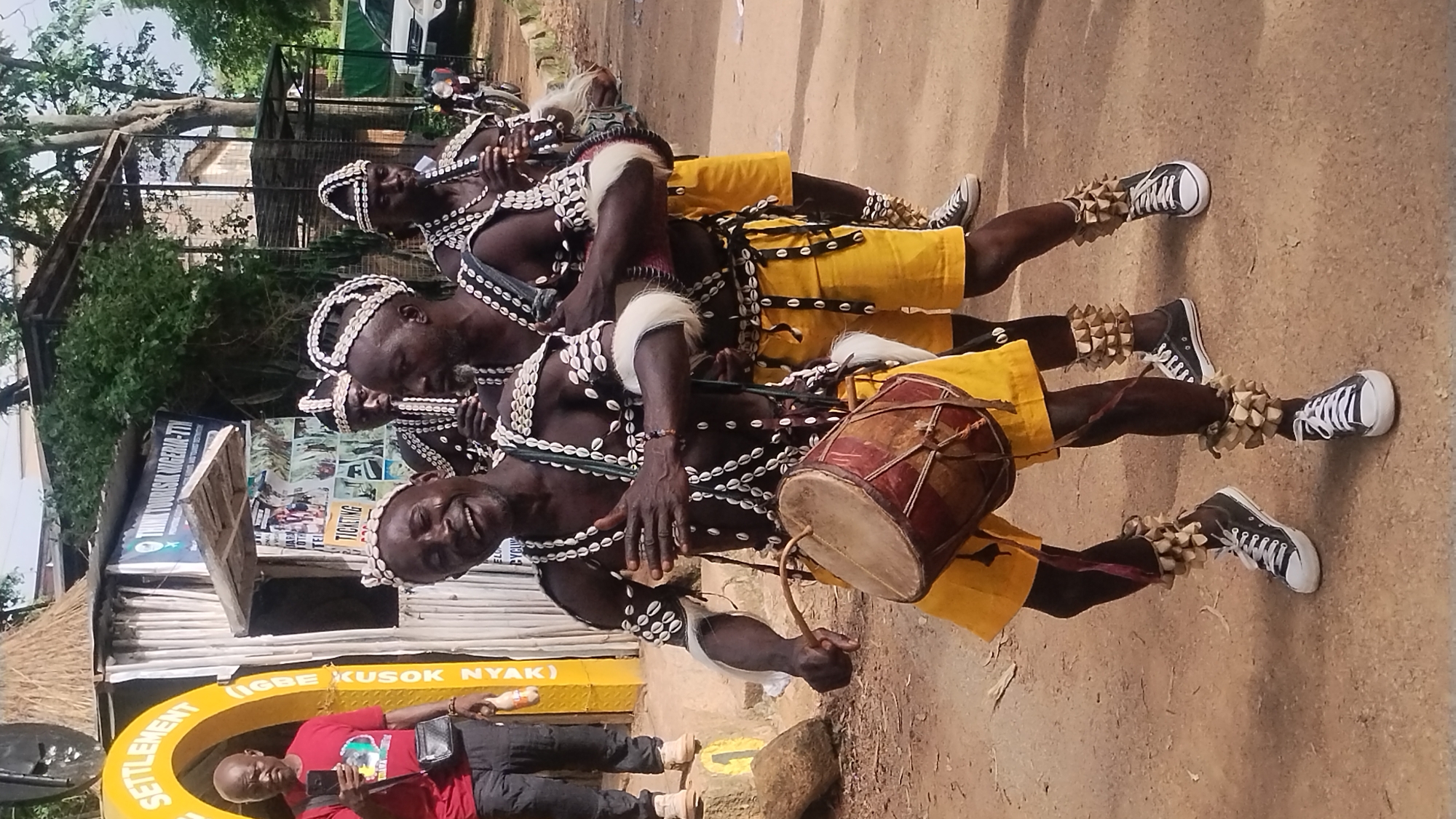 The image depicts a group of four men in traditional attire, likely participating in a cultural event or performance. The central figure, in the foreground, is a Black man wearing a headdress adorned with shells, a necklace of shells, and a yellow skirt. He is playing a drum, which is strapped to his body. His expression is joyful, with his mouth open in a wide smile. Two other men, similarly dressed with headdresses and shell adornments, stand behind him, one of them playing a wind instrument resembling a flute. The fourth man, partially visible to the right, also wears similar attire and plays a wind instrument.
The background suggests an outdoor setting, possibly a street or an open area. A yellow archway with text, including "SETTLEMENT" and "LIGBE KUSOK NYAK (NIGERIA)", is visible to the left. Behind the performers, there is a structure made of bamboo and wood, with signage that includes "THINK TOURISM NIGERIA-TTV". The ground appears to be dirt or sand. In the distance, trees and some buildings are visible, along with a white vehicle and a motorcycle parked on the right side. The lighting indicates it is daytime. The image seems to capture a moment of lively cultural expression. The presence of "Jos, Nigeria" in the prompt suggests this event is taking place in that city.