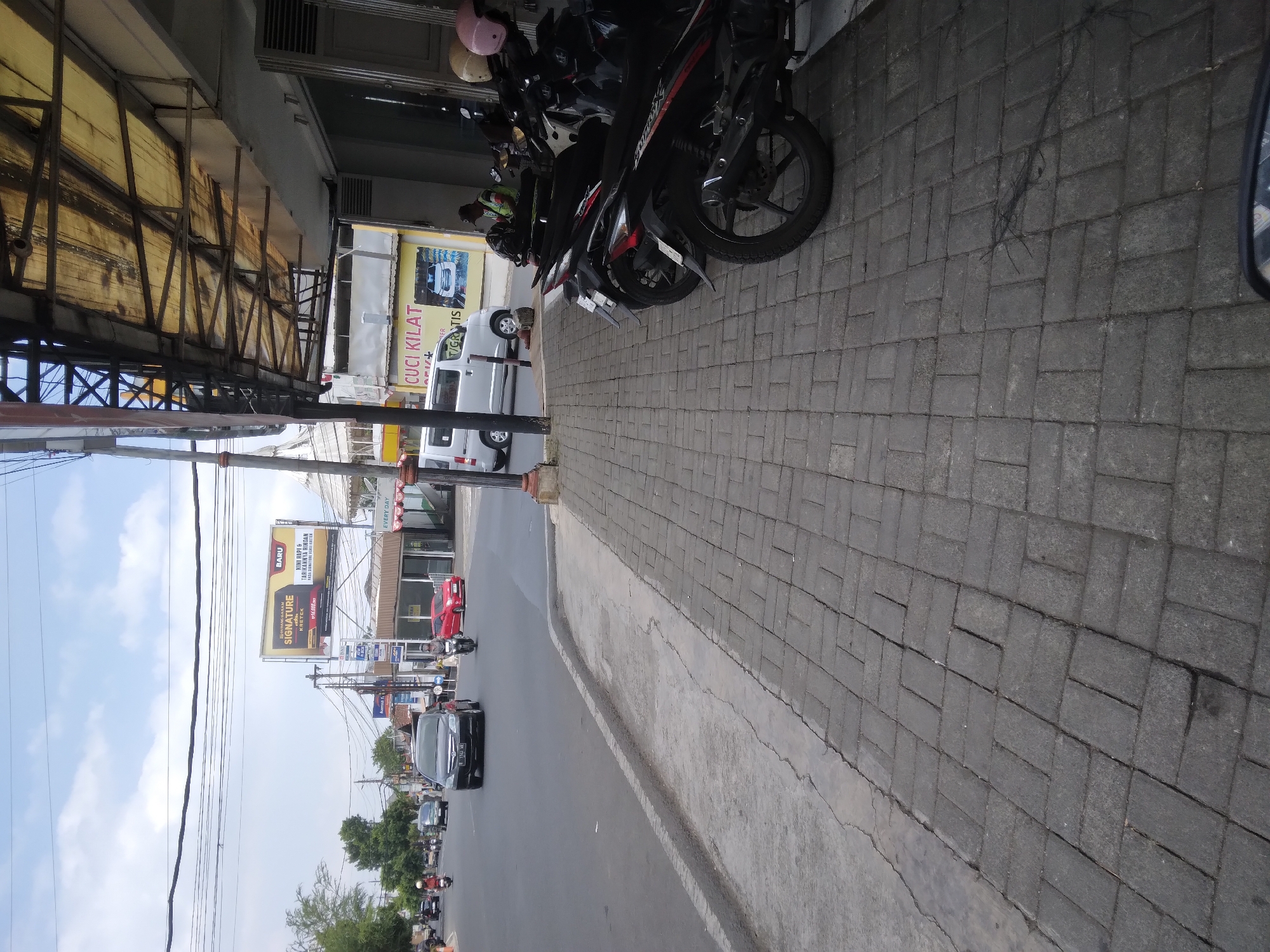 The image depicts a street scene in Berkoh, Indonesia. In the foreground, a grey paved sidewalk runs alongside a road. Several motorcycles are parked on the sidewalk, some with helmets on the handlebars. A white van is visible on the road, as is a red car and a black car. A billboard advertising "Gudang Garam Signature" is prominently displayed in the background. The sky is partly cloudy. The scene appears to be a typical street in an urban or suburban area.