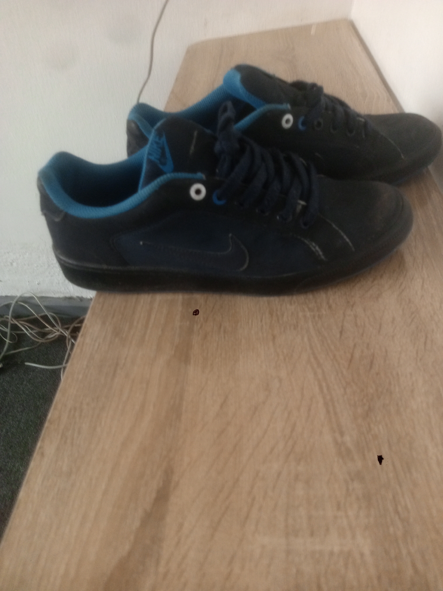 On a light brown, wooden surface, two dark-colored shoes with blue lining are positioned. The shoes appear to be sneakers, laced, and showing signs of wear. The scene is indoors, next to a wall with some electrical wires visible on the left. The location is indiscernible, and there are no people present in the image. The specific location in the city of Abuja, Nigeria, cannot be determined from the provided visual information.
