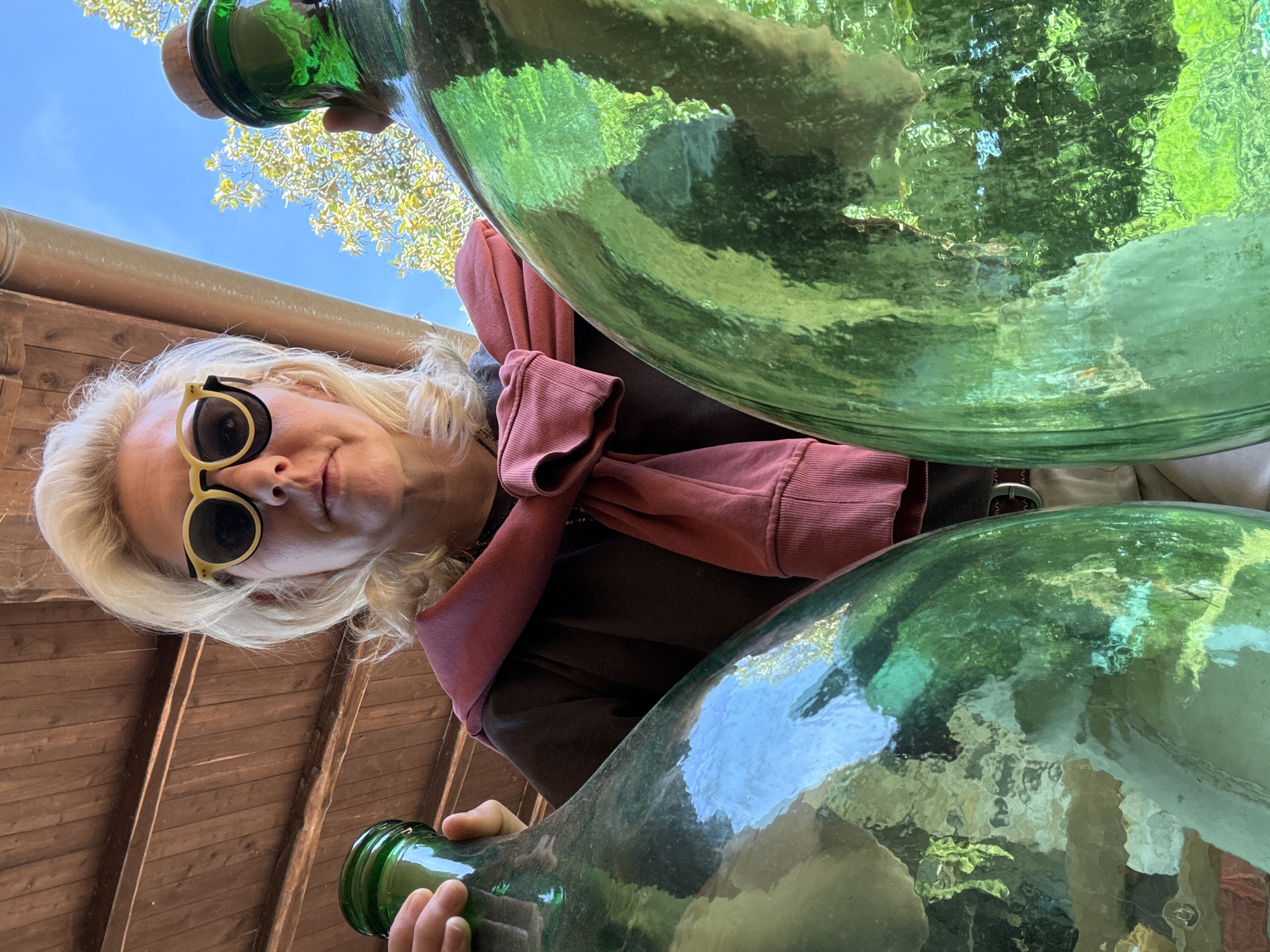 A woman with blonde hair and yellow-rimmed sunglasses looks down at the camera from an unconventional angle, partially obscured by two large, green glass containers that appear to be empty. She wears a brown shirt and a red sweater tied around her shoulders. Her hand is visible holding the neck of one of the glass containers. The sky and tree branches are visible through the glass. Wooden structures are visible in the background, suggestive of a building's exterior. Based on the provided information, this image was taken in the city of Velletri, Italy.