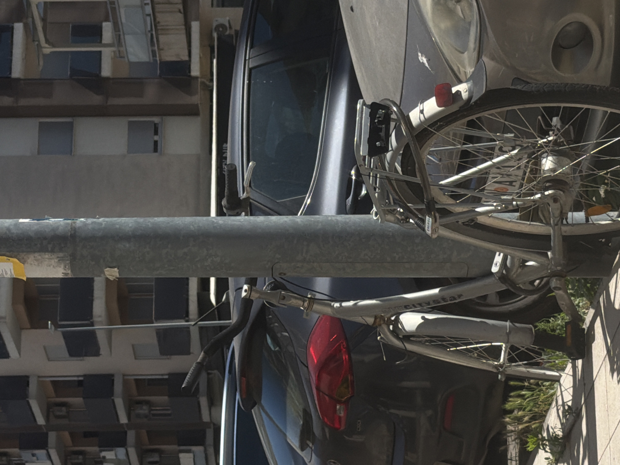 The image depicts a bicycle attached to a metal bar with a car parked beside it. The location appears to be an urban area with buildings in the background. The red tail light of the car is visible. The bicycle is silver with a black tire. A city in Montenegro is not identifiable.
