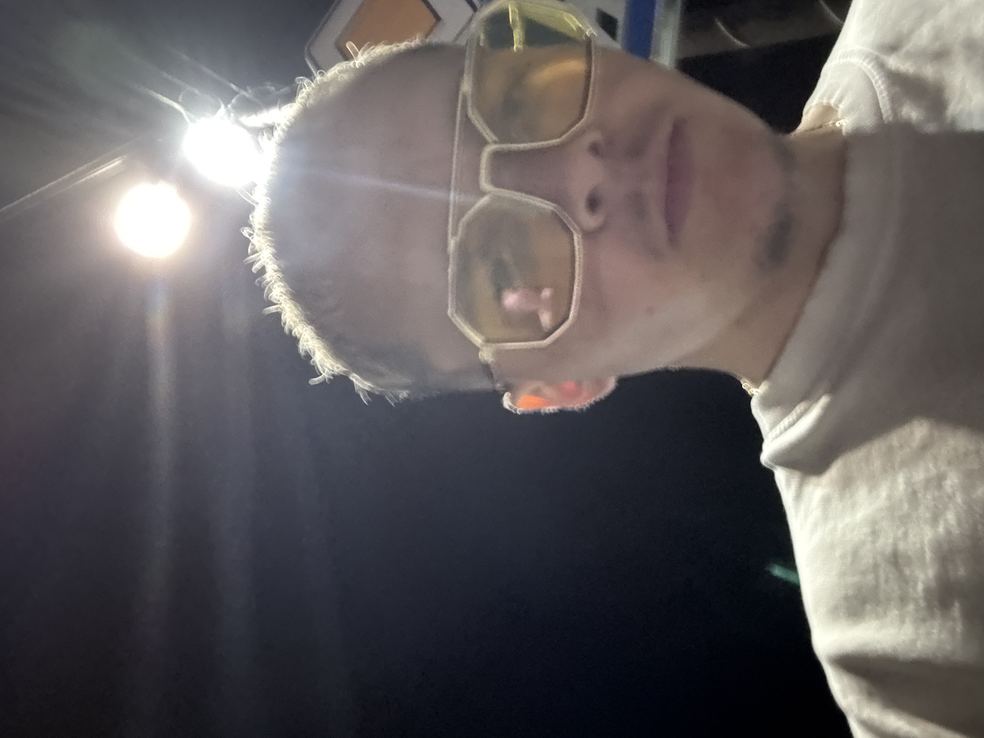 The image is a low-angle, close-up selfie of a person, likely male, taken at night in Kostinbrod, Bulgaria. The subject is positioned off-center to the right, looking downwards into the camera. They have very short, light-colored hair or stubble and wear prominent, oversized yellow-tinted glasses that obscure their eyes. Visible facial hair extends along their jawline and chin. The person is wearing a light-colored, possibly white or off-white, sweatshirt or hoodie, with part of the garment visible on the right side of the frame.
A strong, bright artificial light source, likely a street lamp, is present in the upper left corner, causing significant glare and lens flare. This light creates a halo effect around the subject's head and the rims of their glasses. The background is largely dark, emphasizing the nighttime setting. The person's expression appears neutral. No readable text is visible anywhere in the image.
