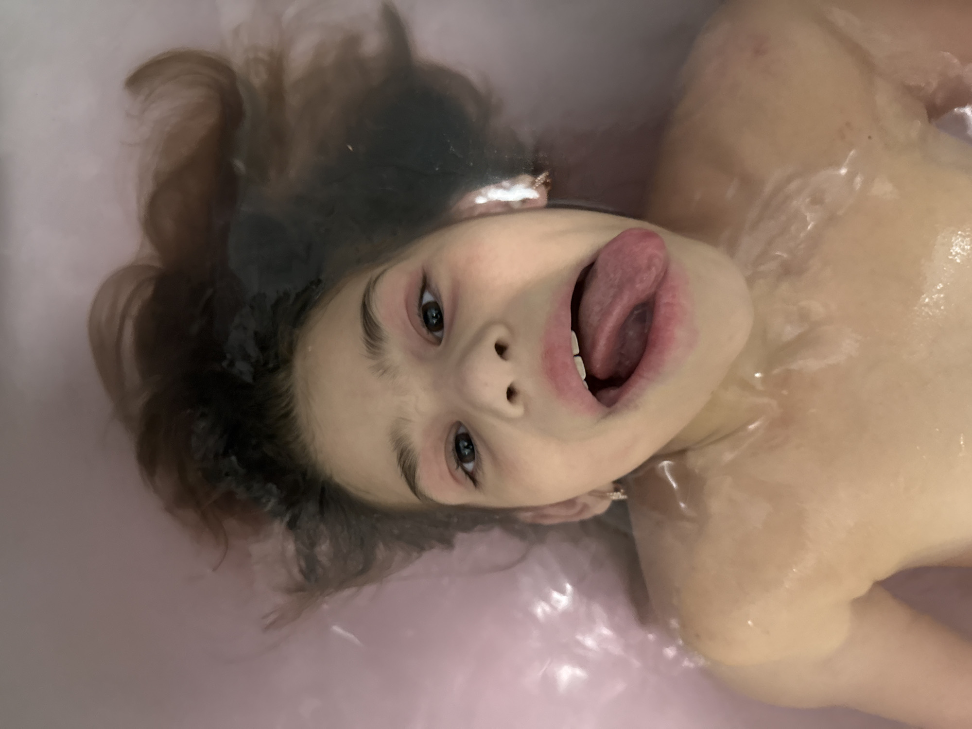 An overhead close-up shot captures a young person, likely a child or pre-teen, immersed in pale pink or lavender-tinted bathwater. The subject's head is centered, and they look directly up at the camera with wide-open eyes. A playful or silly expression is evident, with their mouth open and tongue fully extended. Dark hair is fanned out around their head, partially submerged and wet from the water. The skin of their face and visible upper torso appears wet, with reflections and ripples on the water's surface. The setting is an indoor bathroom, depicting a lighthearted moment during a bath. No text is visible in the image.