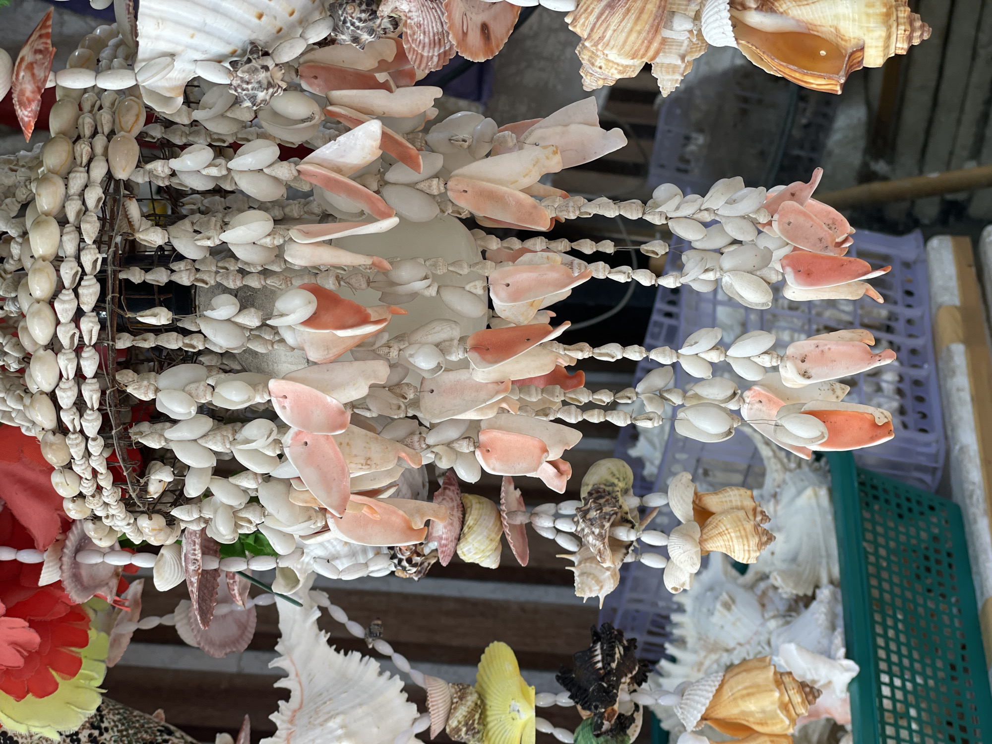The image shows a collection of seashell crafts and decorations hanging from above. The crafts feature various types of seashells, strung together on strings or attached to other shells. The predominant colors are white, beige, and light pink. The background includes glimpses of plastic crates and other indistinct objects. The image was taken in Rawai, Thailand.