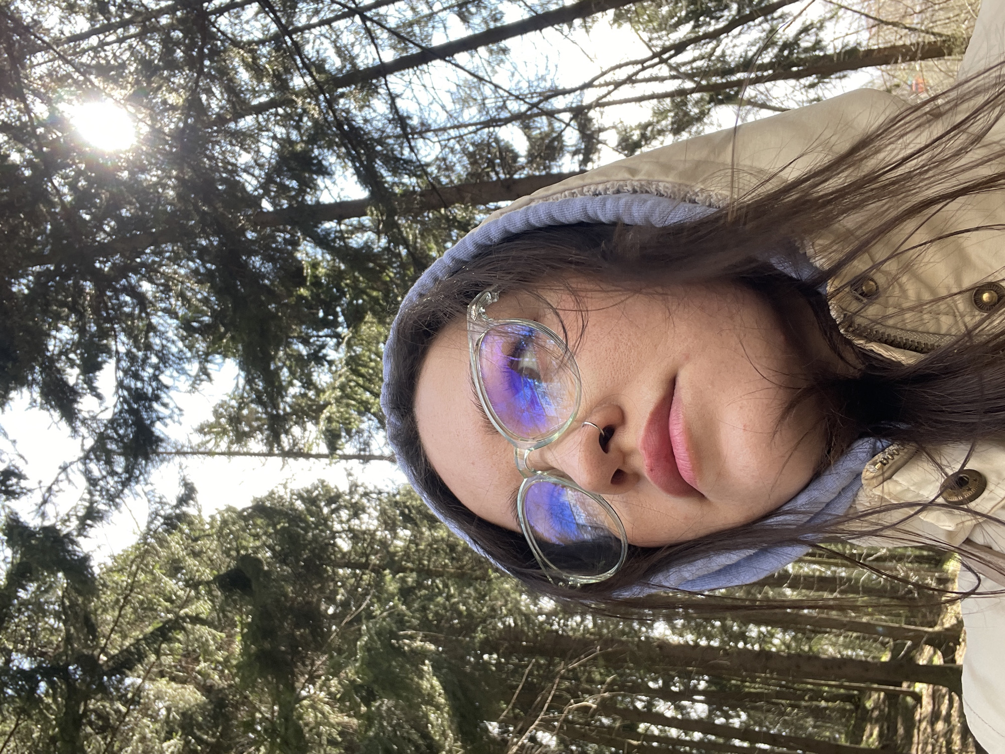 A diagonally oriented selfie features a person looking upwards at a forest canopy. They wear a beige jacket and a blue hoodie. Clear-framed glasses with highly reflective lenses, showing blue and purple tones, are worn over their eyes. Dark hair frames their face, which bears a neutral expression and features a nose ring. Bright sunlight filters through dense evergreen trees in the background, creating a sun flare in the upper left. The low-angle perspective looks upwards into the tree branches. The scene depicts a sunny outdoor setting, likely a forest or wooded area, consistent with locations like Izhevsk, Russia, during daytime. No text is visible.
