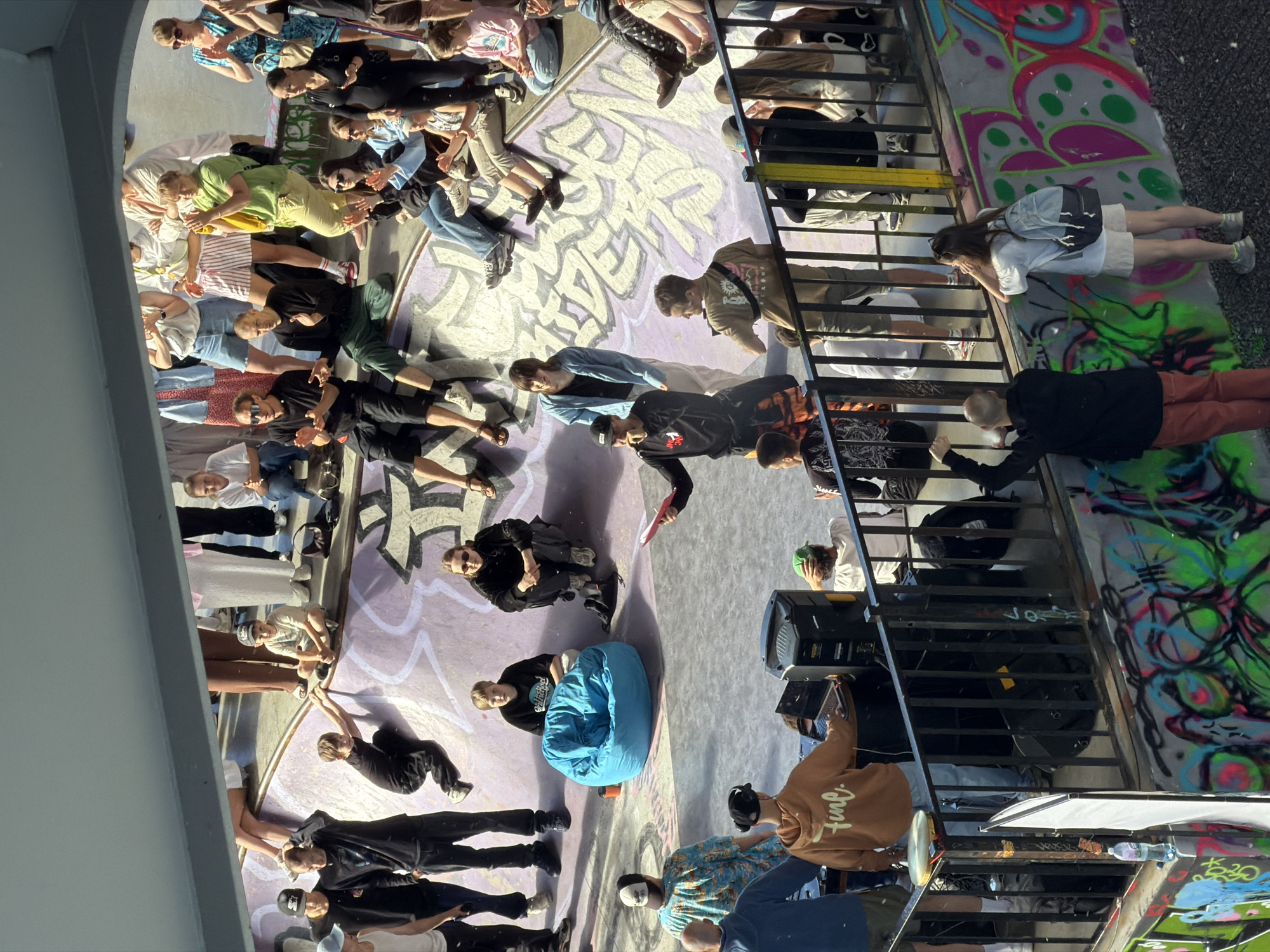 The image depicts a sunny day at a skatepark, likely in Saint Petersburg, Russia, as suggested by the presence of graffiti art that is typical of urban environments in the city. A diverse group of people, including skaters, spectators, and possibly event organizers, are gathered in and around the skatepark.
In the foreground, a person is operating a laptop connected to audio equipment, suggesting a DJ or sound technician at work. Beside them, another person stands with their hand raised, possibly interacting with the DJ or addressing the crowd. A bright orange hoodie with the word "fine." written on it is visible on one individual.
The skatepark itself features concrete ramps with graffiti art adorning the walls. Several individuals are seated on the edges of the ramps, watching the activities. A large, bright blue beanbag chair is placed in the center of the park, offering a casual seating option.
Other individuals are standing, some appear to be skaters, while others are observing. The crowd's attire suggests a casual, outdoor event. The overall atmosphere appears relaxed and energetic, with people enjoying the skatepark activities under the bright sunlight. The presence of the graffiti and the urban setting strongly indicate a location within Saint Petersburg.