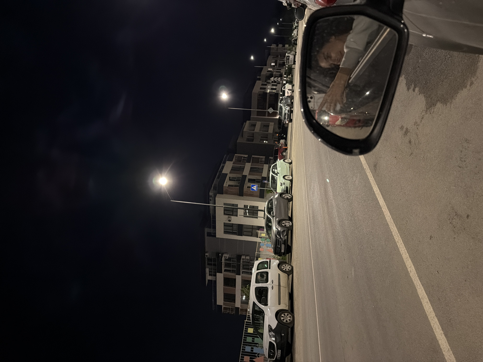 A nighttime exterior scene depicts a street in Sofia, Bulgaria, with a row of modern apartment buildings lining the right side of the road. Several cars are parked along the sidewalk, including a white van, a silver SUV, and a light green compact car. A street lamp illuminates the area, casting a bright light on the road and pavement. In the foreground, a car's side mirror reflects a young woman leaning out of the passenger window, taking a selfie with her phone. The reflection shows her smiling and her arm extended. The road is asphalt with white lane markings. The sky is dark, with a hint of moonlight or distant light sources visible.