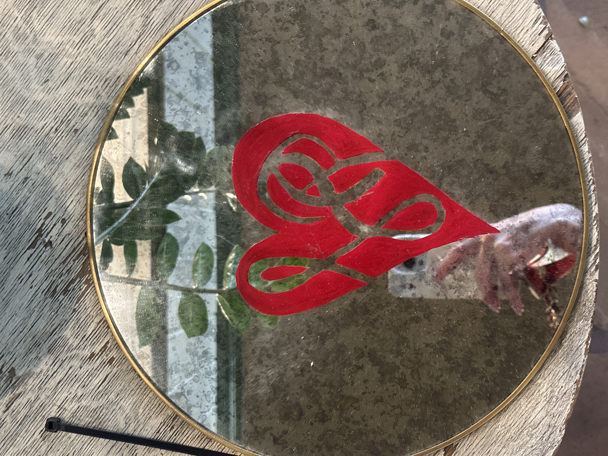 A round mirror with a thin gold frame sits on a distressed wooden surface. A red, heart-shaped design is affixed to the mirror's surface. The reflection in the mirror reveals a leafy plant, a window, and a hand holding a smartphone, likely capturing the image. A black zip tie is present on the wooden surface next to the mirror. The location is unknown.