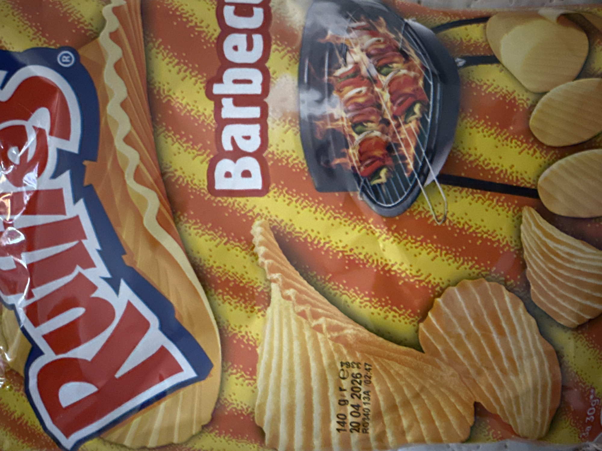A forensic, matter-of-fact description of the image:
The image is a close-up shot of a bag of "Ruffles" brand potato chips, barbecue flavor. The package is predominantly yellow and orange, with a stylized illustration of potato chips, shish kabobs on a grill, and the word "Barbecue" in white block letters. The expiration date "20 04 2026" and weight of "140 gr" are printed on the package. The setting or location cannot be determined from the image, nor can a location in Varna, Bulgaria be assessed.