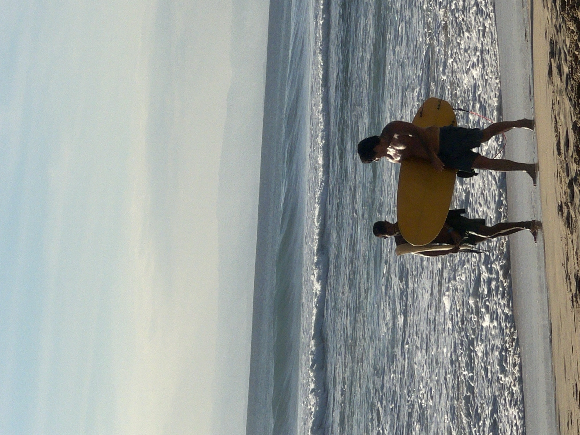 Two men are standing on the beach near the ocean in Kuta Selatan, Indonesia. One man is carrying a yellow surfboard, and both men are wearing shorts. The sky is cloudy, and the ocean is calm.