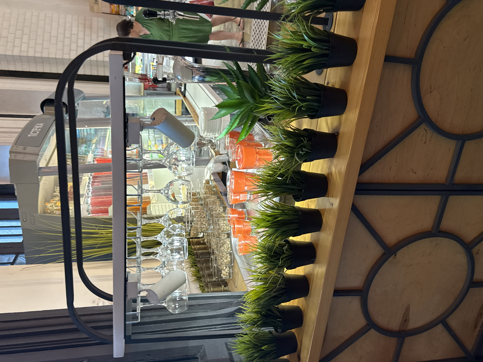 The image shows a shelf unit with several rows of potted plants and glassware. The plants appear to be artificial, with green, spiky leaves. The glassware includes wine glasses and tumblers, some filled with orange liquid. In the background, a person wearing a green shirt is visible, standing behind a counter with what appears to be a beverage dispenser. The setting seems to be indoors, possibly a cafe or bar, with brickwork visible in the upper left corner. The overall impression is one of a retail or service environment, with decorative elements like the plants and the circular design on the wooden shelf. There is a sign with Cyrillic text that reads "ПІСТЕЦКА" visible on the left side of the frame, likely indicating a brand or product.