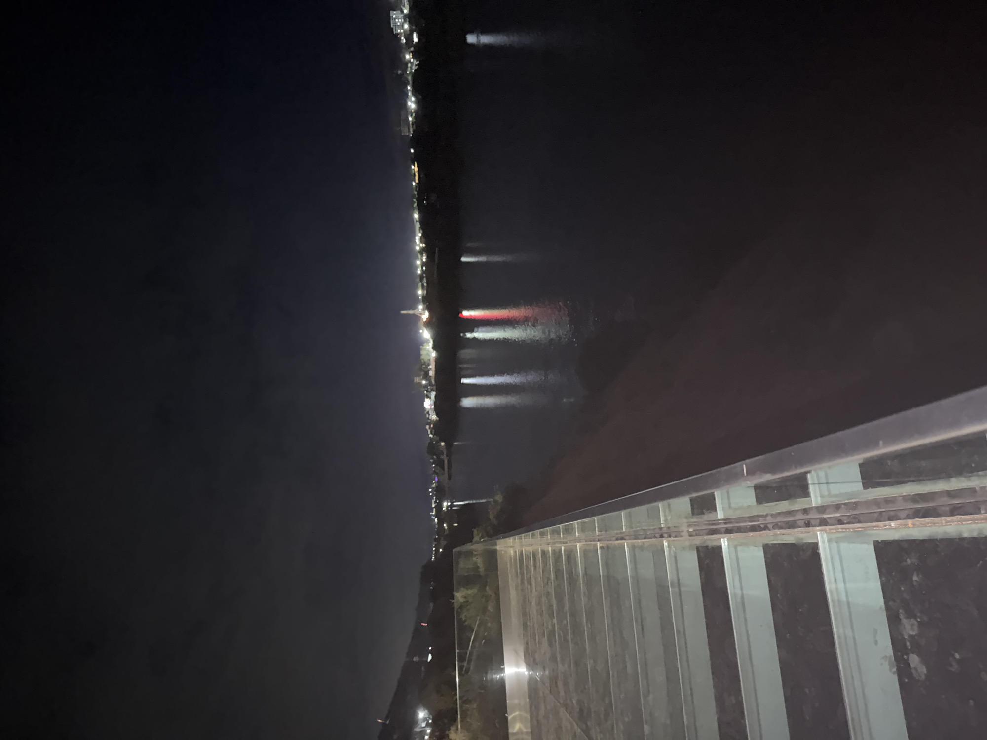The image depicts a nighttime scene from a glass-floored walkway overlooking a wide, dark body of water, likely a river or lake, with a city skyline in the distance. The walkway itself is composed of thick glass panels supported by metal beams, offering a view downwards. On the left side of the walkway, there are some green plants and a railing.
The cityscape across the water is illuminated by numerous lights, suggesting a populated area. A prominent, tall structure, possibly a tower or a bridge pylon, is visible amongst the city lights, emitting a bright light at its peak. There are reflections of these lights shimmering on the surface of the water. The sky above is dark and cloudy. The overall impression is that of a modern scenic viewpoint in an urban environment at night. The location is identified as Chinarkent, Uzbekistan.