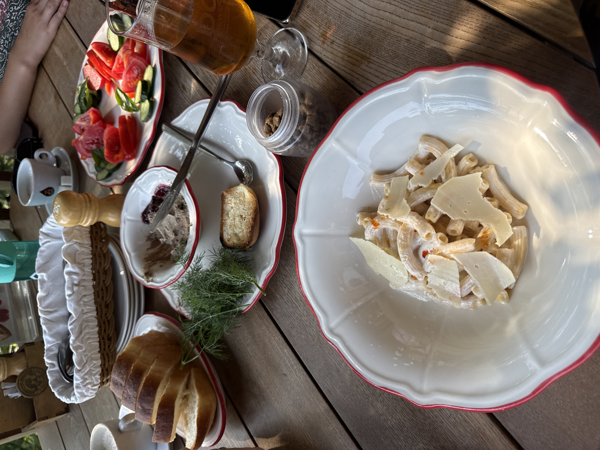 An overhead, eye-level shot captures a table laden with food on a wooden surface. In the foreground, a white bowl with a red rim contains pasta topped with shaved cheese and red specks, possibly chili flakes. To the left, sliced bread sits on a plate. Behind it, a white basket holds silverware. Further back, a small white bowl contains a spread, likely pâté, with a spoon resting on it. Next to that, a plate displays sliced tomatoes and cucumbers. A glass of amber liquid, possibly beer, sits to the right. A small glass jar filled with dark, granular material is placed near the glass. In the background, a cup and saucer are visible, along with a salt shaker and a green bottle. Sunlight casts shadows across the table. The scene suggests a meal being enjoyed outdoors, possibly in the city of Progress, Russia, though this location cannot be definitively confirmed from the visual data alone.