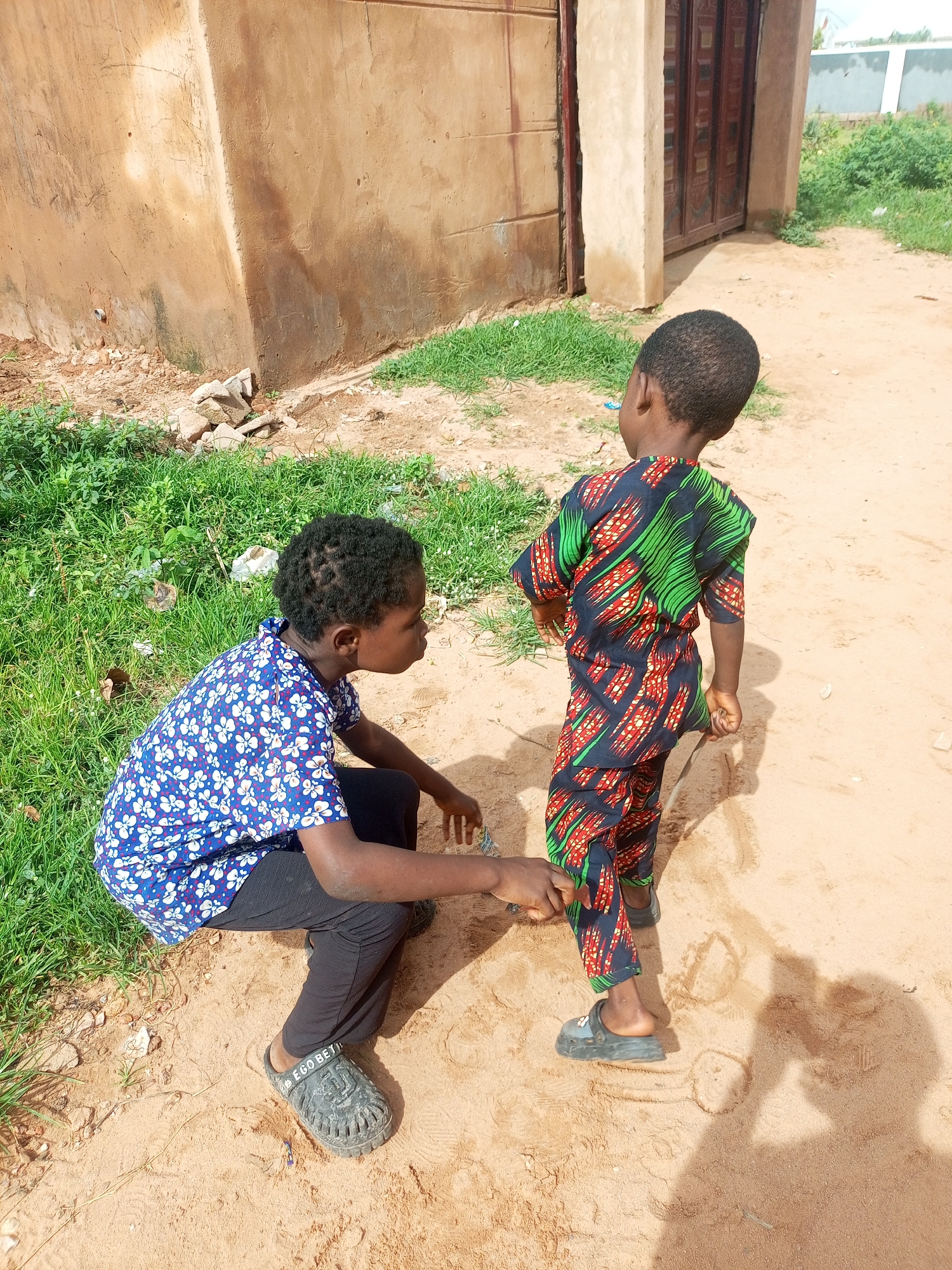 In Tempure, Nigeria, two children are playing outside on a sandy surface next to a tan-colored building with a large, dark wooden door. One child, viewed from the side and slightly from behind, kneels in the sand. This child wears a blue shirt with a white floral pattern and black pants, along with gray clogs. The other child stands facing away from the camera, wearing a dark blue, green, and red patterned outfit, also in dark clogs. The ground is a mix of sand and sparse green grass, with some debris visible. Sunlight casts shadows on the sandy area.