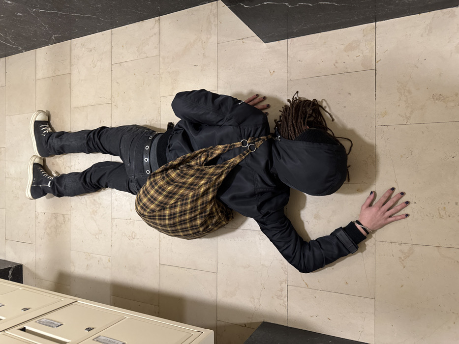 A person lies prone on a tiled floor, wearing black jeans, a black jacket with a hood, and high-top sneakers. A large, plaid bag rests on their back. Their dreadlocks spill out from under the hood. Their hands are extended, fingers spread, revealing dark nail polish. A mailbox bank is in the lower left corner of the frame.