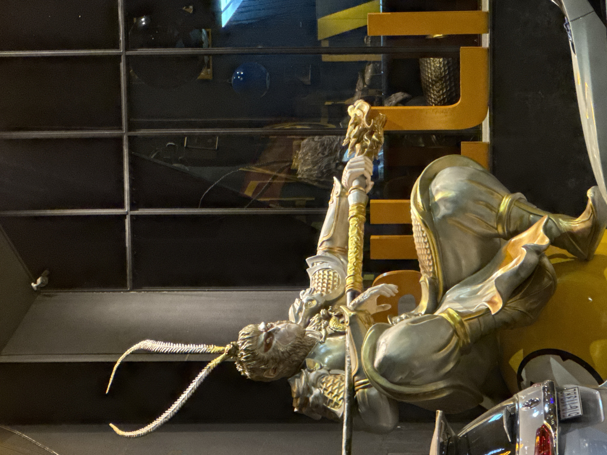 The image depicts a statue of the Monkey King, Sun Wukong, seated and holding a staff. The statue is elaborately detailed, with golden armor and a monkey-like face. It is positioned in front of a window display, with large, bright orange letters spelling out "LI" visible behind it. In the foreground, the rear of a silver car with a visible license plate is present. The scene appears to be outdoors, likely in an urban setting, possibly a shop or an entertainment venue in Da Lat, Vietnam. The lighting suggests it is nighttime or dusk.
