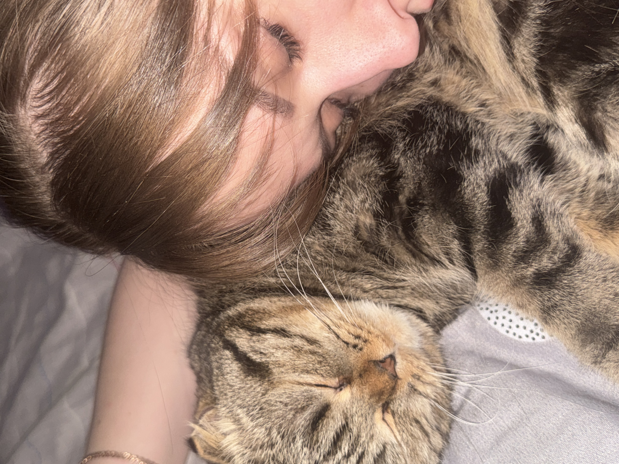A close-up shot features a person and a tabby cat lying close together, both with their eyes closed. The person, positioned on the left, has brown hair and is lying on their side, with their face partially pressed against the cat. The cat, a brown tabby, rests its head towards the bottom of the frame, its face visible with eyes shut. The two are lying on a light gray surface, possibly a bed or blanket. No identifiable location in the city of Chaikovsky, Russia is present.