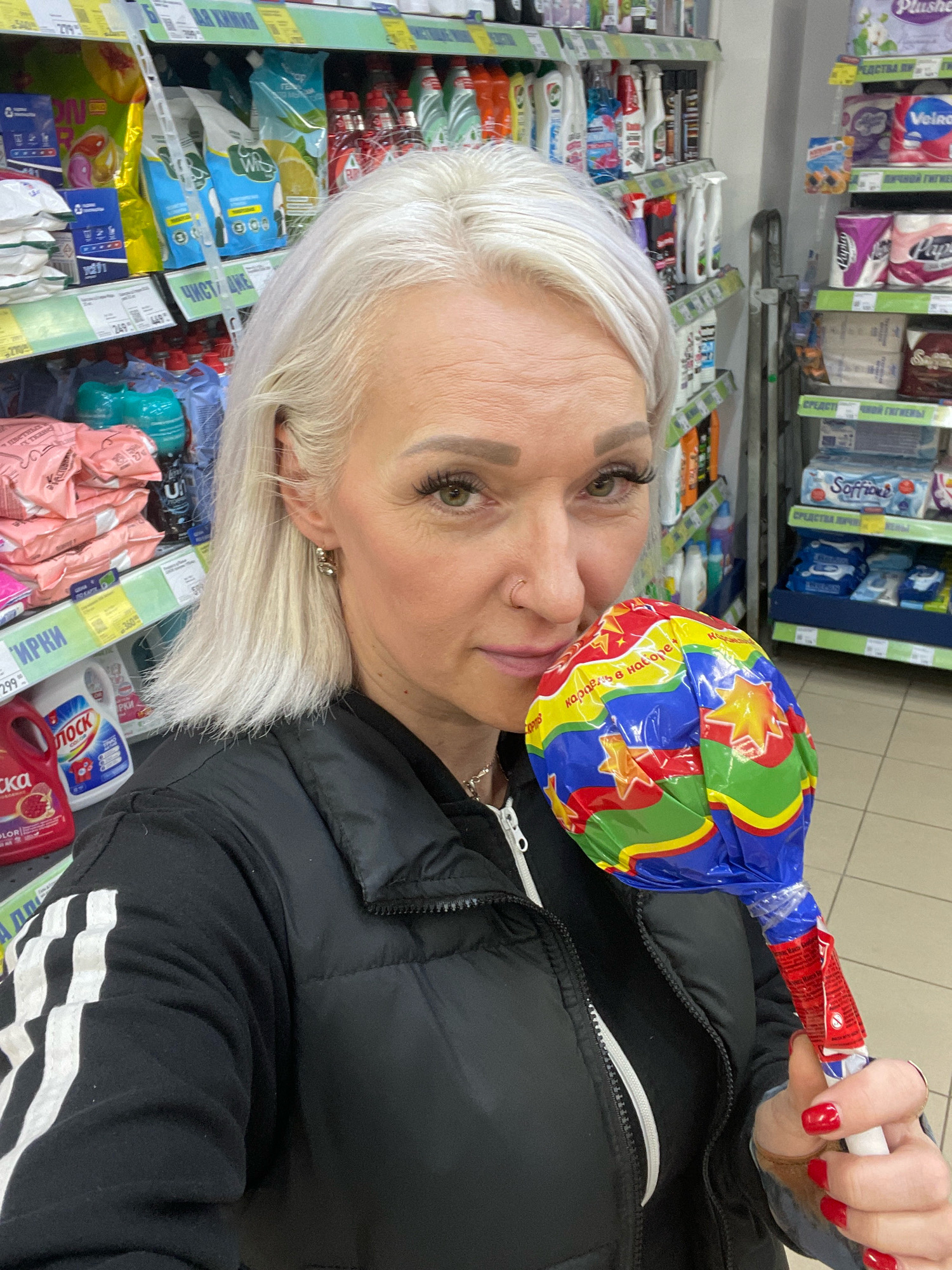 The image shows a woman with blonde hair holding a large, colorful lollipop. She is wearing a black puffer jacket with white stripes on the sleeves and a silver necklace. The background appears to be a supermarket aisle, with shelves stocked with various products, including cleaning supplies and toiletries. Some of the visible text on the shelves and products is in Russian. Based on the visible Cyrillic text, the location is likely a supermarket in Saint Petersburg, Russia. The woman is looking at the camera and smiling slightly, with her right hand holding the lollipop.