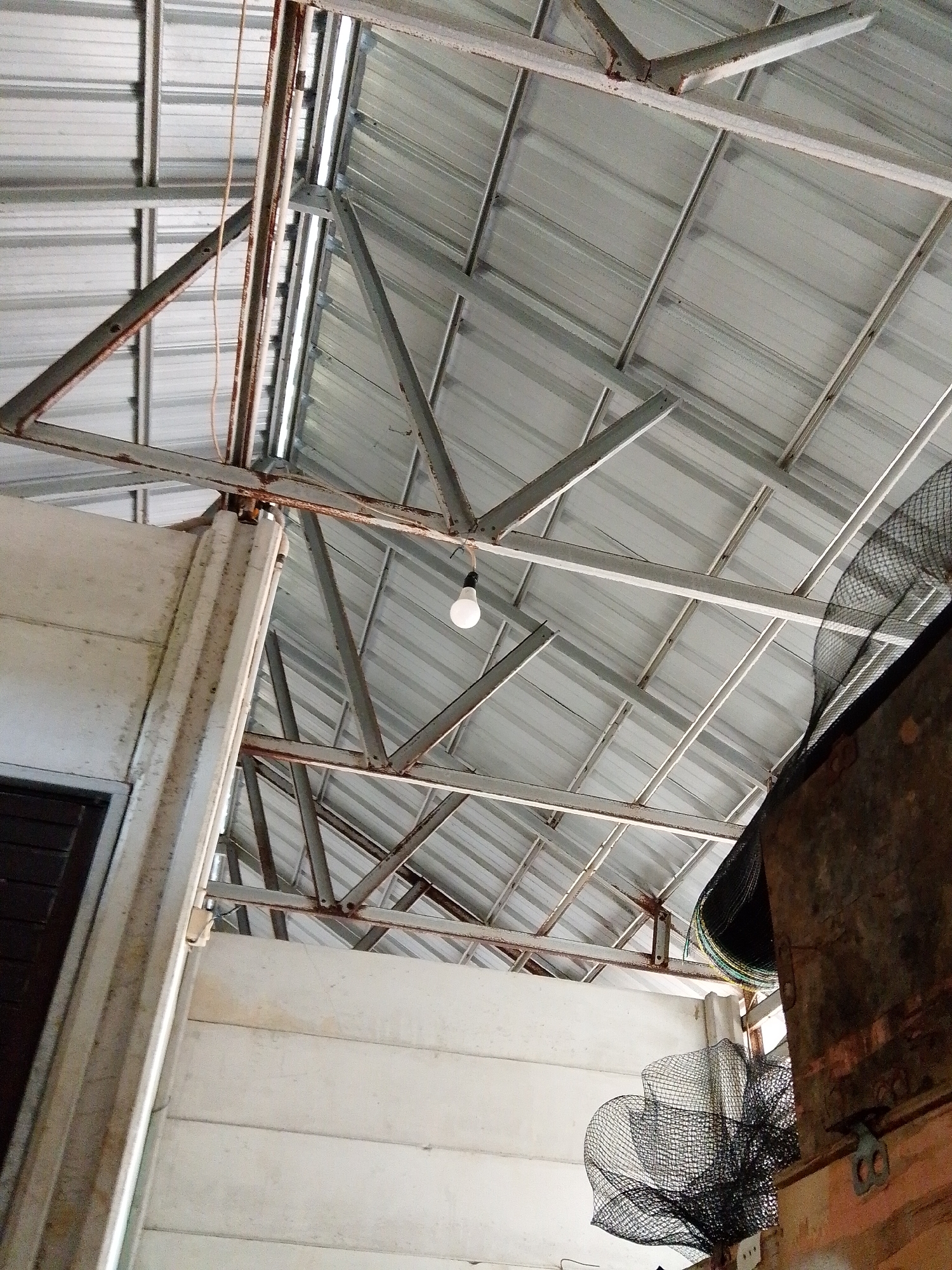 The image shows an interior view of a structure, likely a building, located in Bukit Ketok, Indonesia. The perspective is looking upwards towards a metal roof supported by a framework of metal beams, some showing signs of rust. A single light bulb hangs from the frame. Sections of the walls are visible, constructed with a white, plank-like material. Additionally, a black net-like fabric is draped along part of the right side. A dark, wooden or metal fixture is visible on the right-hand side of the image. The scene appears to be utilitarian.
