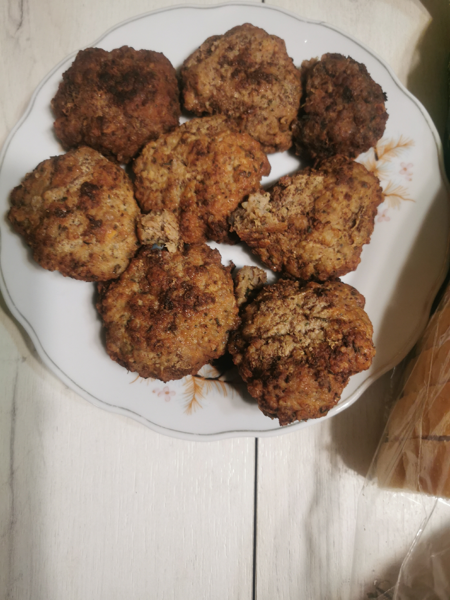 A high-angle, medium shot depicts a white porcelain plate with a floral design, holding eight fried patties. The patties vary in color from light brown to dark brown. The plate sits on a white wooden surface with vertical planks. To the right of the plate, there is a partial view of what appears to be bread in a transparent plastic bag. The location is not discernible from the image, and there is no evidence to suggest the location is Stara Zagora, Bulgaria. No people, actions, or interactions are visible in the image.