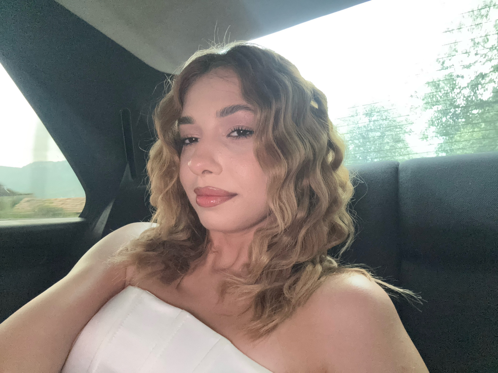 The image shows a young woman with wavy blonde hair, wearing a white strapless top. She is seated in the back of a car, with the window on her left showing a blurred view of trees and buildings. The light from the window illuminates her face and shoulder. The interior of the car appears to be dark fabric. The scene suggests a journey, possibly through the city of Ijevan, Armenia, given the glimpses of trees and structures visible outside the window. The overall mood is casual and relaxed.