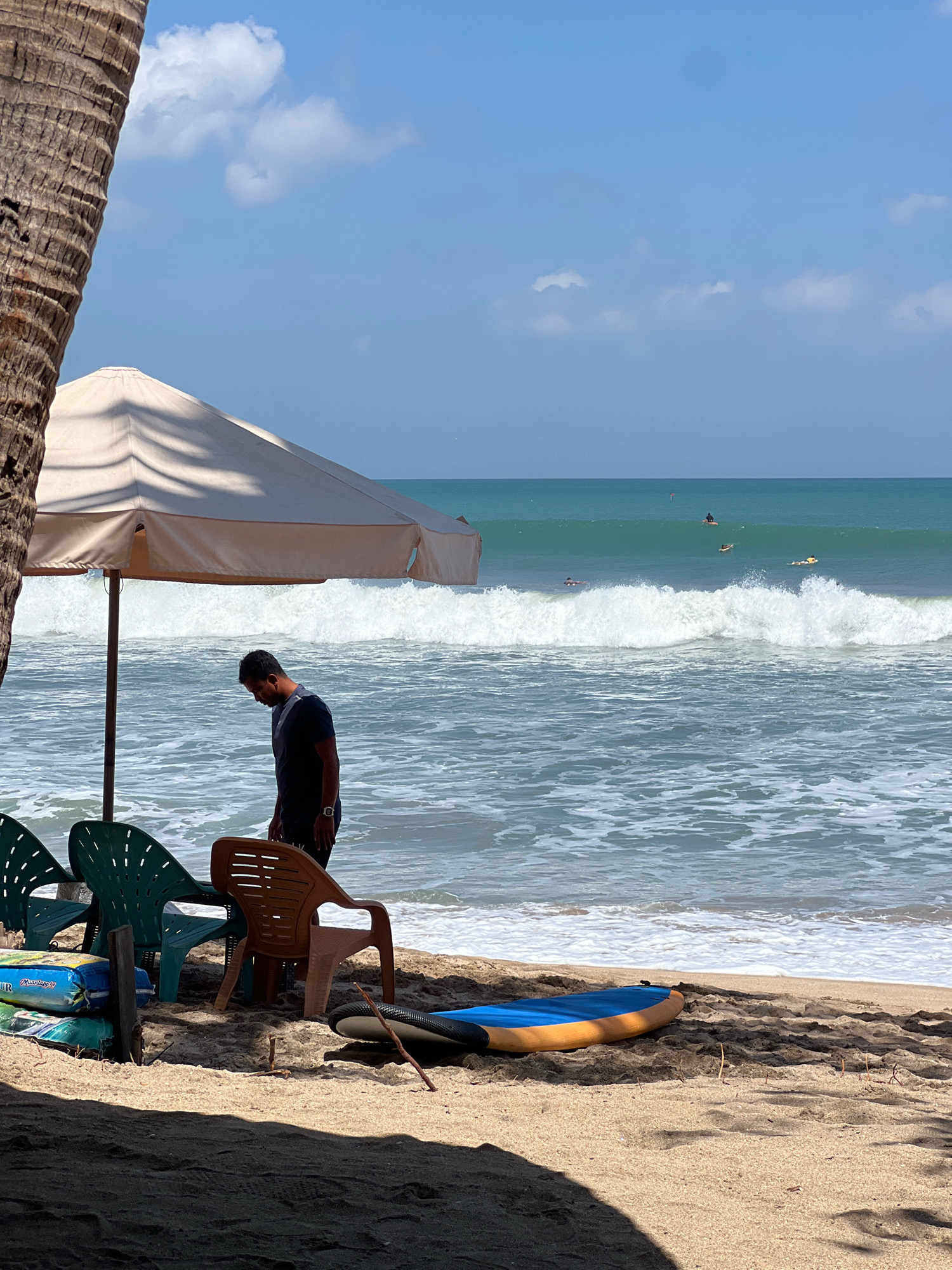 A man stands under a large umbrella on a sandy beach in Kuta, Indonesia, looking down. The ocean is calm with small waves, and several surfers are visible in the distance. Beach chairs and surfboards are scattered on the sand. A palm tree trunk is on the left side of the frame. The sky is blue with some white clouds.