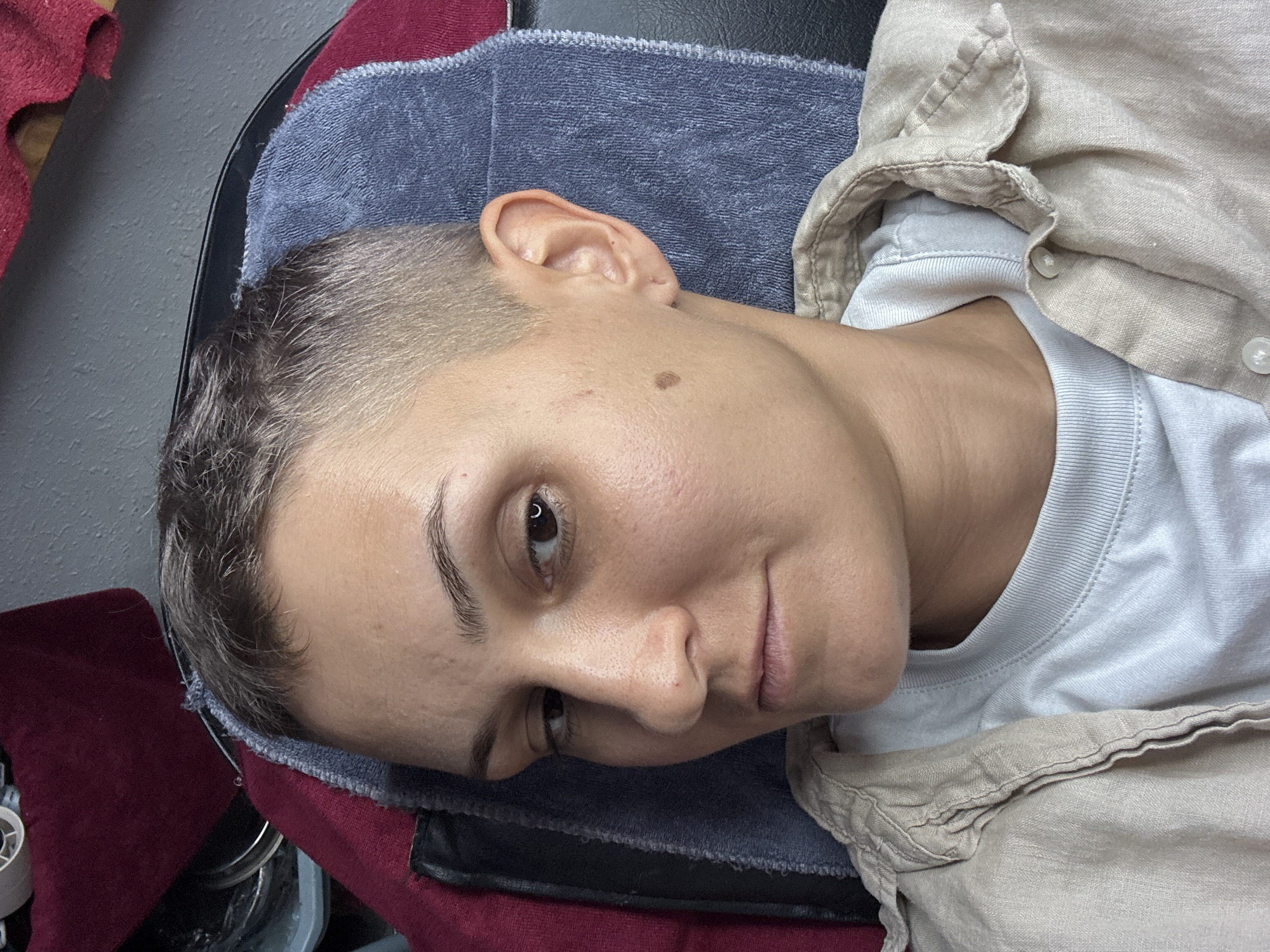 A close-up, high-angle shot shows a person with very short, dark hair, styled in a buzz cut. The person is wearing a light-colored t-shirt underneath an unbuttoned, light tan linen shirt. The background features a dark gray wall and a dark red fabric draped over something to the left. A blue towel or fabric is visible behind the person's head and neck. The person is looking towards the left of the frame with a neutral expression. A mole is visible on the right cheek. The scene does not provide any indicators of a specific location within Nha Trang Ward, Vietnam.