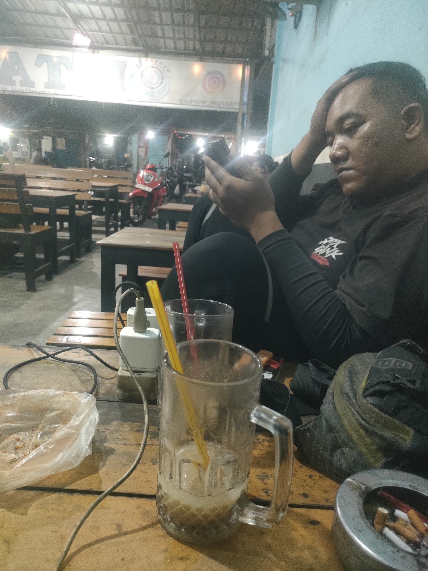 A medium shot captures a man in a black long-sleeved shirt sitting at a wooden table in an outdoor food stall or restaurant in Jogosatru, Indonesia. The man is holding a smartphone in his right hand and resting his left hand on his forehead, appearing to be in thought or perhaps experiencing discomfort. In front of him on the table are several items: a glass mug with ice and a beverage, a plastic bag containing food remnants, a white electrical outlet with a charging cable, and an ashtray filled with cigarette butts. In the background, a red motorcycle is visible, along with rows of wooden tables and chairs, and the exterior of a building with a sign displaying the letters "AT" prominently. The lighting suggests it is nighttime.