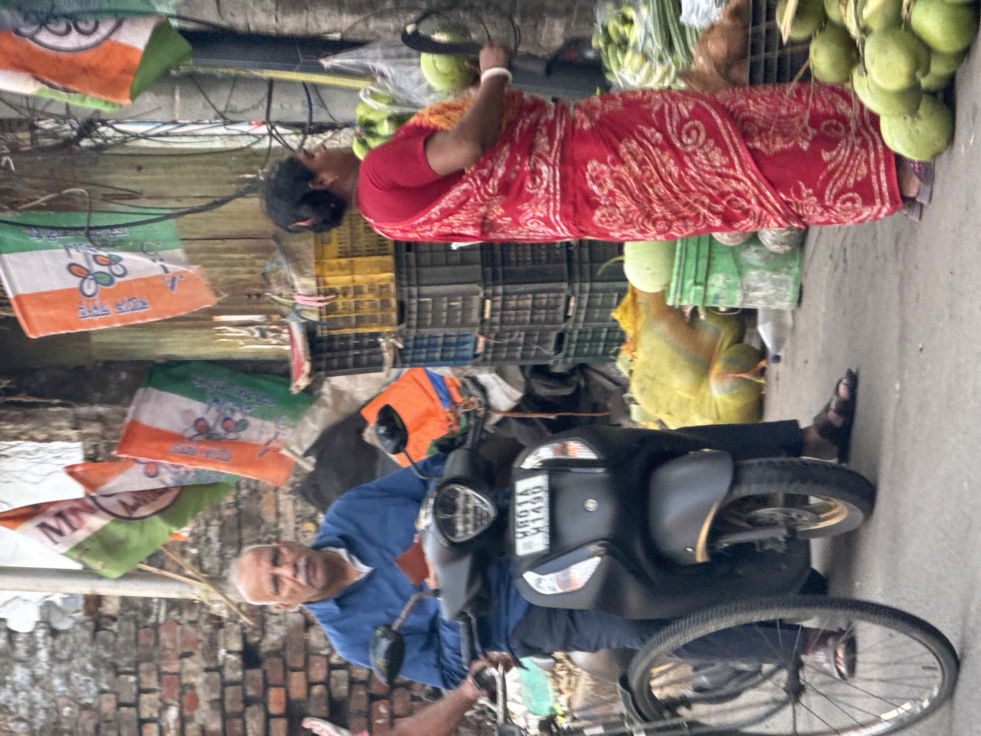 A street scene in Kolkata, India, shows an elderly man sitting on a black scooter, dressed in a blue shirt and dark pants, partially obscured by a bicycle propped against the scooter. A woman in a red and white patterned sari stands nearby. Behind them are crates, bags, and coconuts for sale. Political party flags hang overhead, adding to the cluttered street atmosphere.