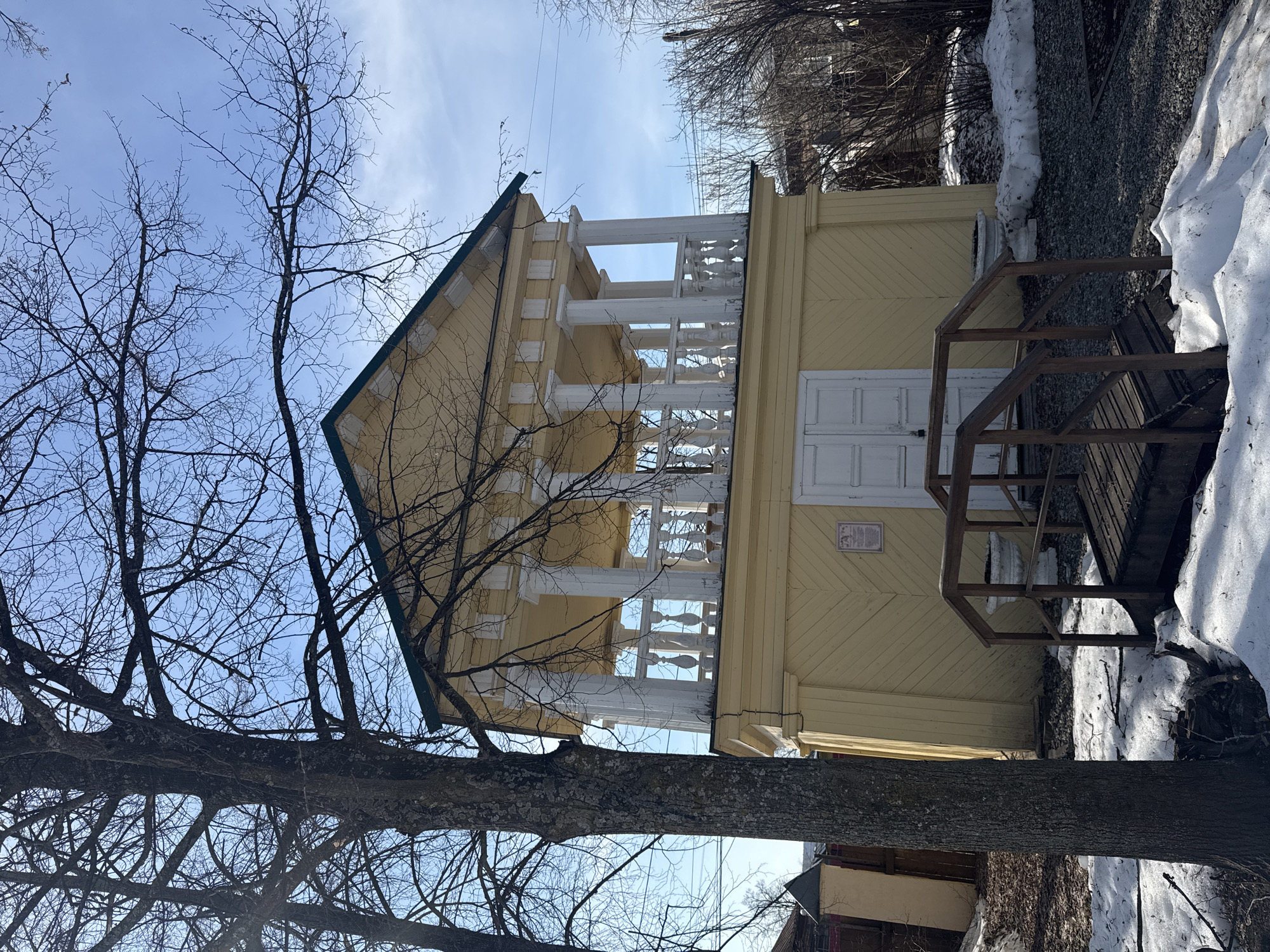 The image shows a two-story yellow wooden house in Votkinsk, Russia. The house has a green roof and white decorative trim around the windows and balconies. A wooden staircase leads to a porch. Patches of snow are visible on the ground next to the house. A bare tree occupies the left side of the frame, partially obscuring the view of the house. The sky is blue with some clouds.