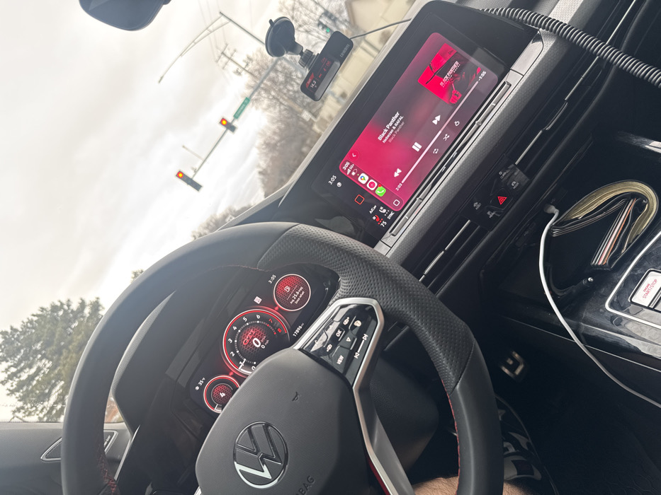 The image depicts the interior perspective of a modern Volkswagen GTI vehicle, stationary at an intersection in Bloomingdale, United States of America. The vehicle is facing an intersection with two traffic lights, both illuminated red. A green street sign, partially visible, indicates "SCHM... RD," consistent with Schmale Road.
The time displayed on the digital instrument cluster and the infotainment system is 3:05. The external temperature is 35 degrees Fahrenheit, suggesting cool or cold weather. The sky is overcast.
Inside the vehicle, a black multi-function steering wheel with red stitching and a central VW logo is prominent. The digital instrument cluster shows "GTI," a speed of 0 MPH, 17,896 miles on the odometer, and an average fuel economy of 25.6 MPG. The infotainment system displays Apple CarPlay, actively playing the song "Black Panther" by Nle Choppa & Bp FAL, with the volume at 75%. A dashcam is mounted on the windshield via a suction cup. A driver's left arm is partially visible on the lower-left side of the frame. A white charging cable is connected to a device in the center console area, near the engine start/stop button and a hazard light button.
Outside the vehicle, through the windshield, bare deciduous trees and some evergreen foliage are visible, indicative of a dormant season. Utility poles and overhead wires are also present.