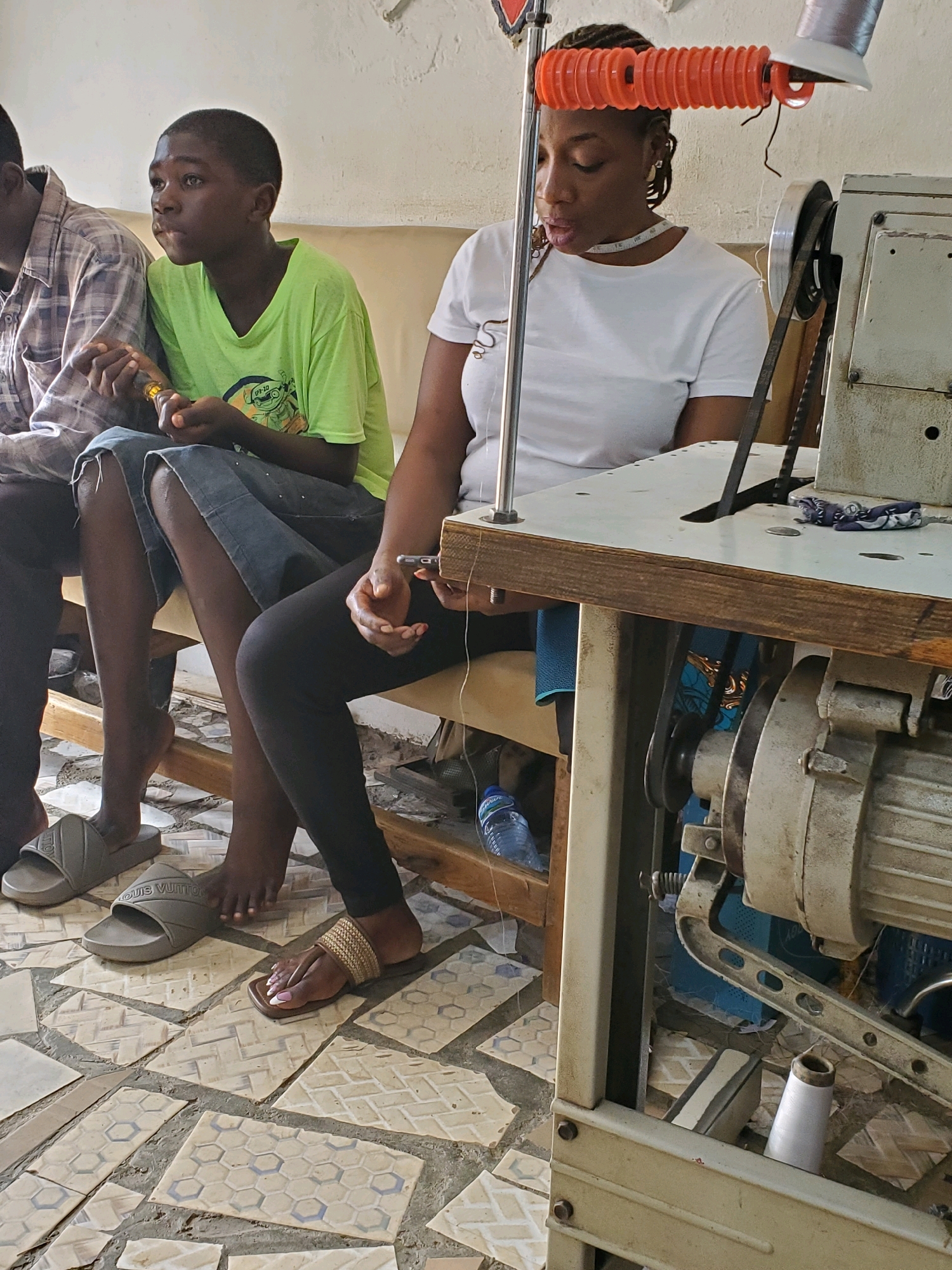 A medium shot captures a sewing workshop in Jos, Nigeria, with three individuals seated on a beige upholstered bench. On the left, a young boy, possibly a teenager, wears a lime green t-shirt and denim shorts, holding a yellow object. Beside him, a woman with braided hair is dressed in a white t-shirt and black leggings, actively engaged with a sewing machine. Her right hand holds a mobile phone. The sewing machine, a substantial industrial model, is positioned on a wooden table. In the background, another person, partially visible, wears a plaid shirt. The floor is tiled with a pattern of beige and white geometric shapes. A blue water bottle is visible beneath the bench. The scene is brightly lit.