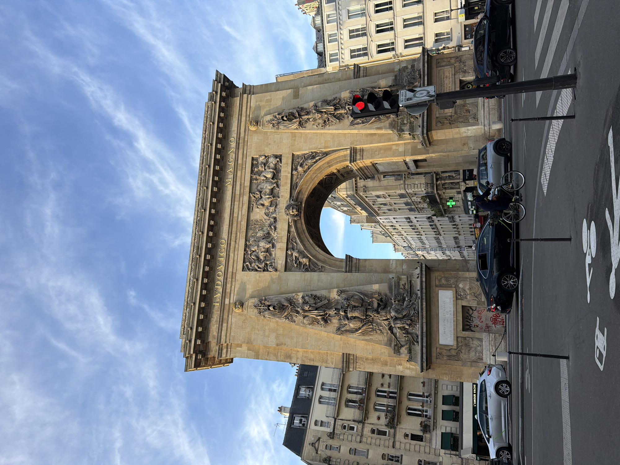 The media file captures an eye-level view of the Porte Saint-Denis, a monumental triumphal arch in Paris, France, during what appears to be late afternoon or early evening. The impressive arch, constructed from light-colored stone, dominates the scene and is richly adorned with sculptural reliefs depicting military themes and allegorical figures.
Prominently displayed on the arch's frieze are gold-lettered inscriptions: "LUDOVICO" on the left and "MAGNO" on the right, together reading "LUDOVICO MAGNO" (To Louis the Great), dedicated to King Louis XIV. A plaque on the arch features the Latin text "PREFECTUS ET AEDILES ANNO DOMINI MDC LXXII" (Prefects and Aediles in the Year of Our Lord 1672), indicating its dedication date.
The arch spans a busy city street where several vehicles are present, including a black sedan parked partially beneath the arch and a white hatchback further down. A person on a bicycle, wearing a dark jacket, is also visible on the street. A red traffic light is illuminated on a pole attached to the arch, suggesting traffic is stopped. Road markings for a crosswalk and a bicycle lane are painted on the asphalt. A sign attached to the traffic light reads "FIN DE VOIE OUVERTE AUX BUS" (End of lane open to buses). Below the arch, a shopfront displays the name "L'AMOUR VACHE".
Surrounding the arch are typical multi-story Parisian buildings with numerous windows, extending into the background. The sky above is a clear blue with scattered, thin white clouds. The overall scene depicts a normal day in a bustling urban environment, with no specific event taking place beyond the routine flow of city life. The lighting is bright and even, highlighting the architectural details.